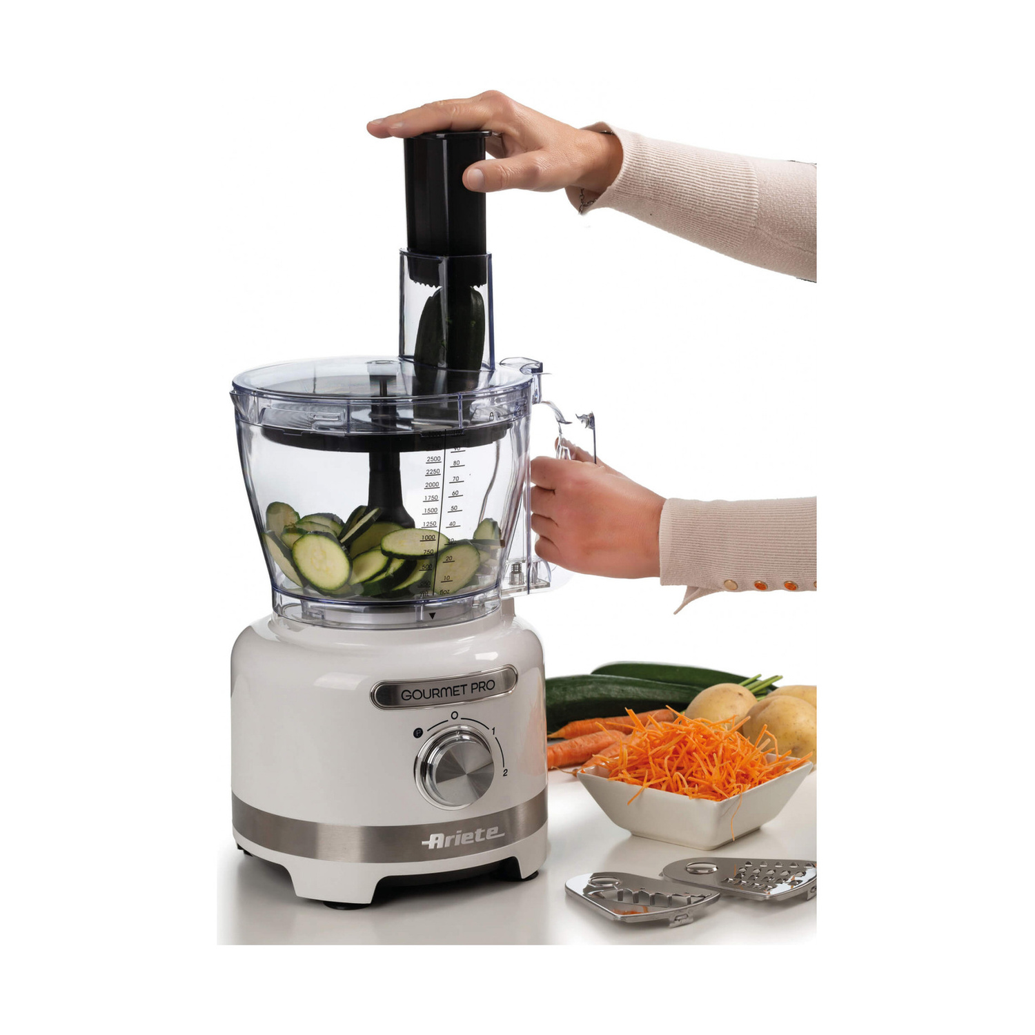 Una mano aziona un robot da cucina bianco Ariete Gourmet Pro con zucchine affettate nella ciotola.