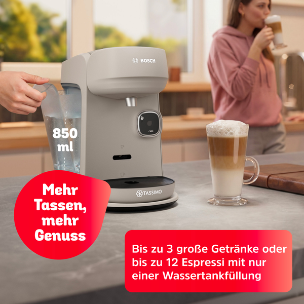 Beige Bosch Tassimo Kaffeemaschine mit Wassertank auf einer Theke, daneben ein Latte Macchiato und eine Frau im Hintergrund.