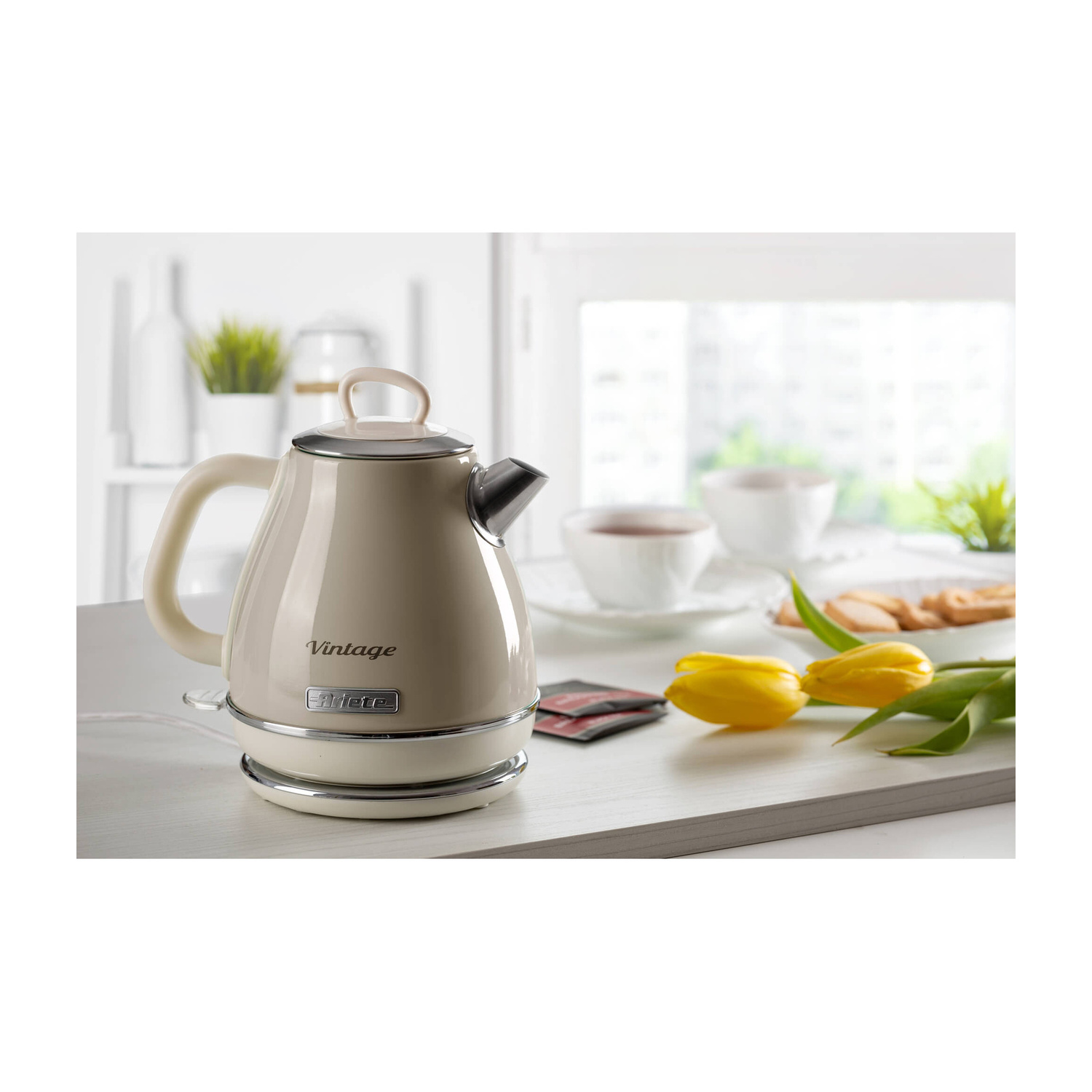 Bollitore retro beige su un bancone accanto a tulipani gialli, tazze e biscotti in una cucina luminosa.