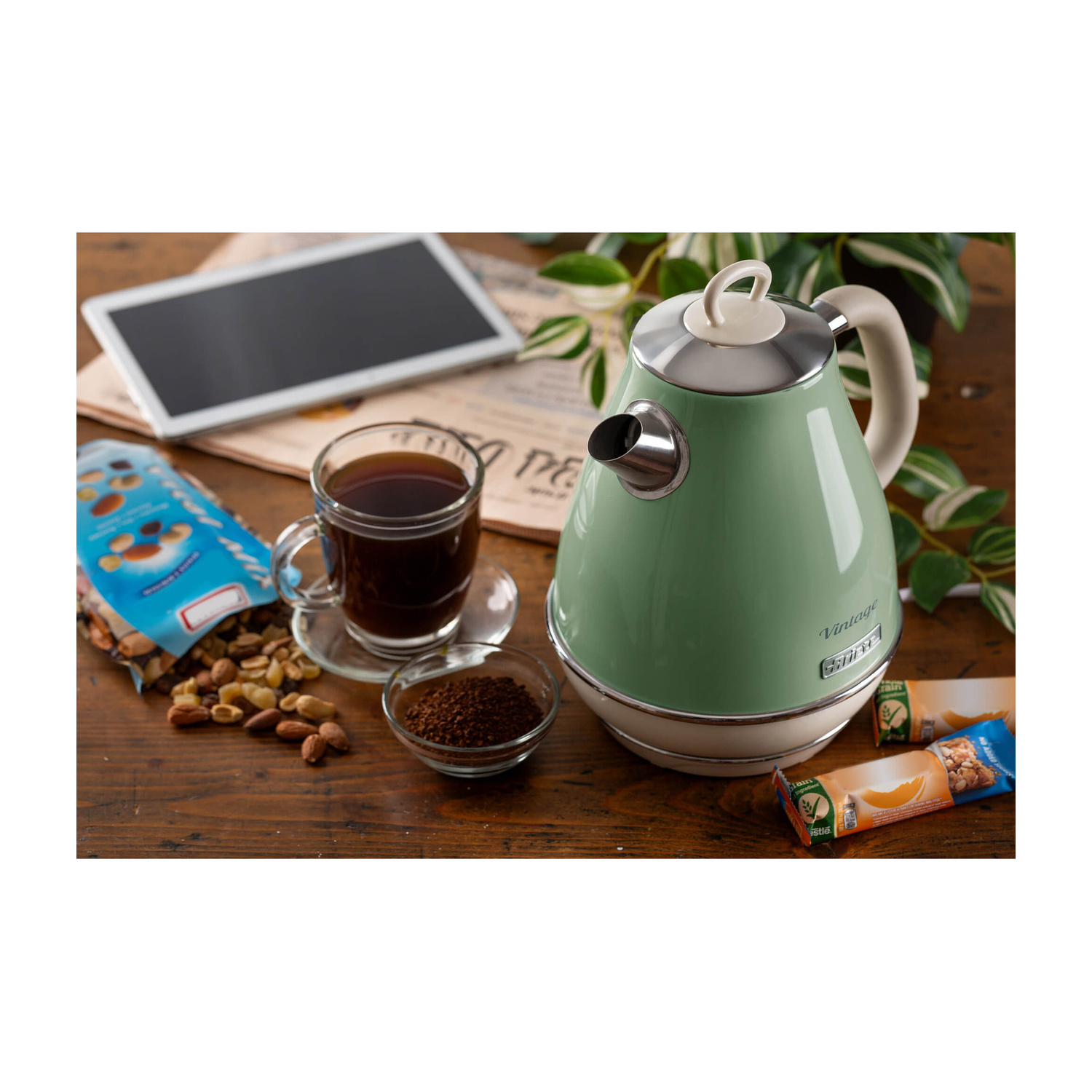 Un bollitore verde, una tazza di caffè, noci, un sacchetto di cereali e un tablet su un tavolo di legno.