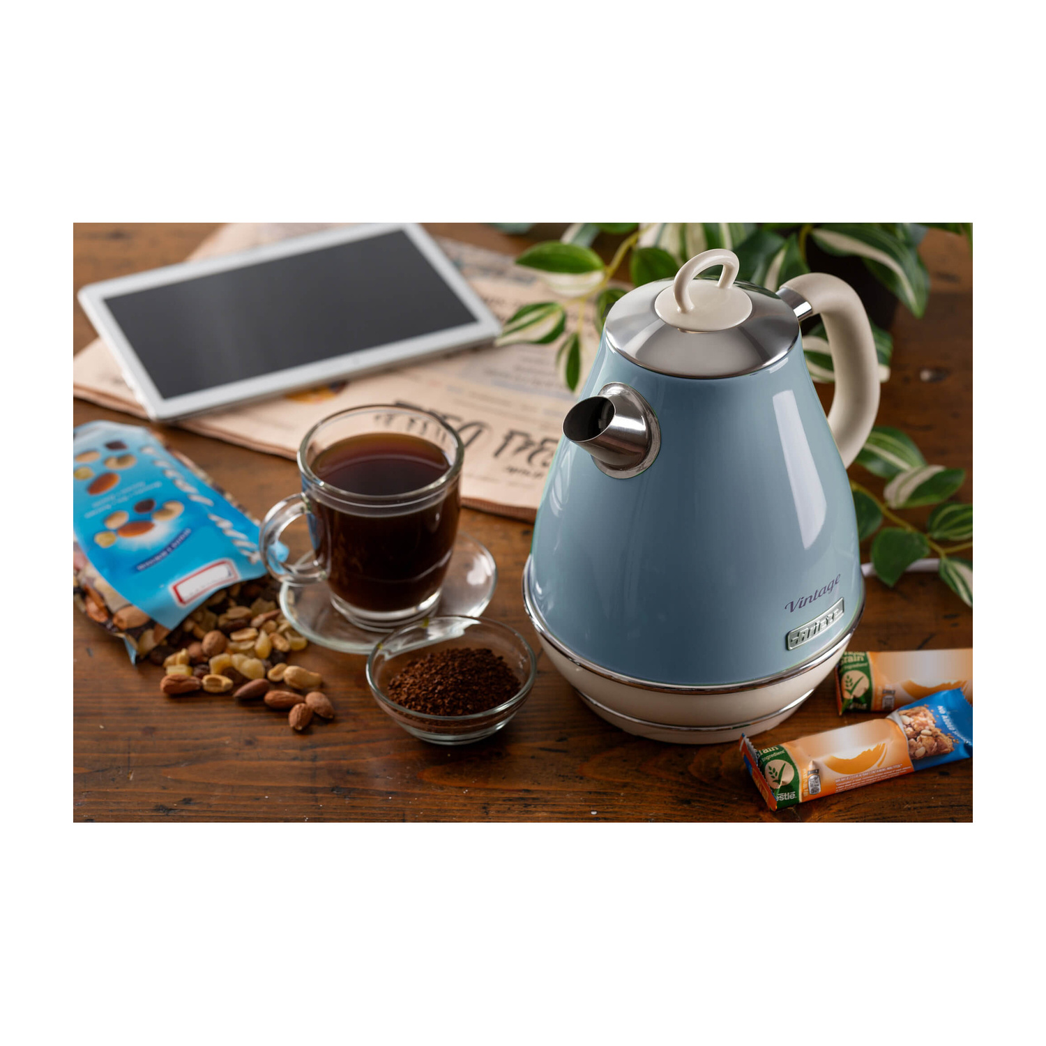 Un bollitore vintage blu, tazza di caffè, tablet, frutta secca e barrette di cereali su un tavolo di legno.