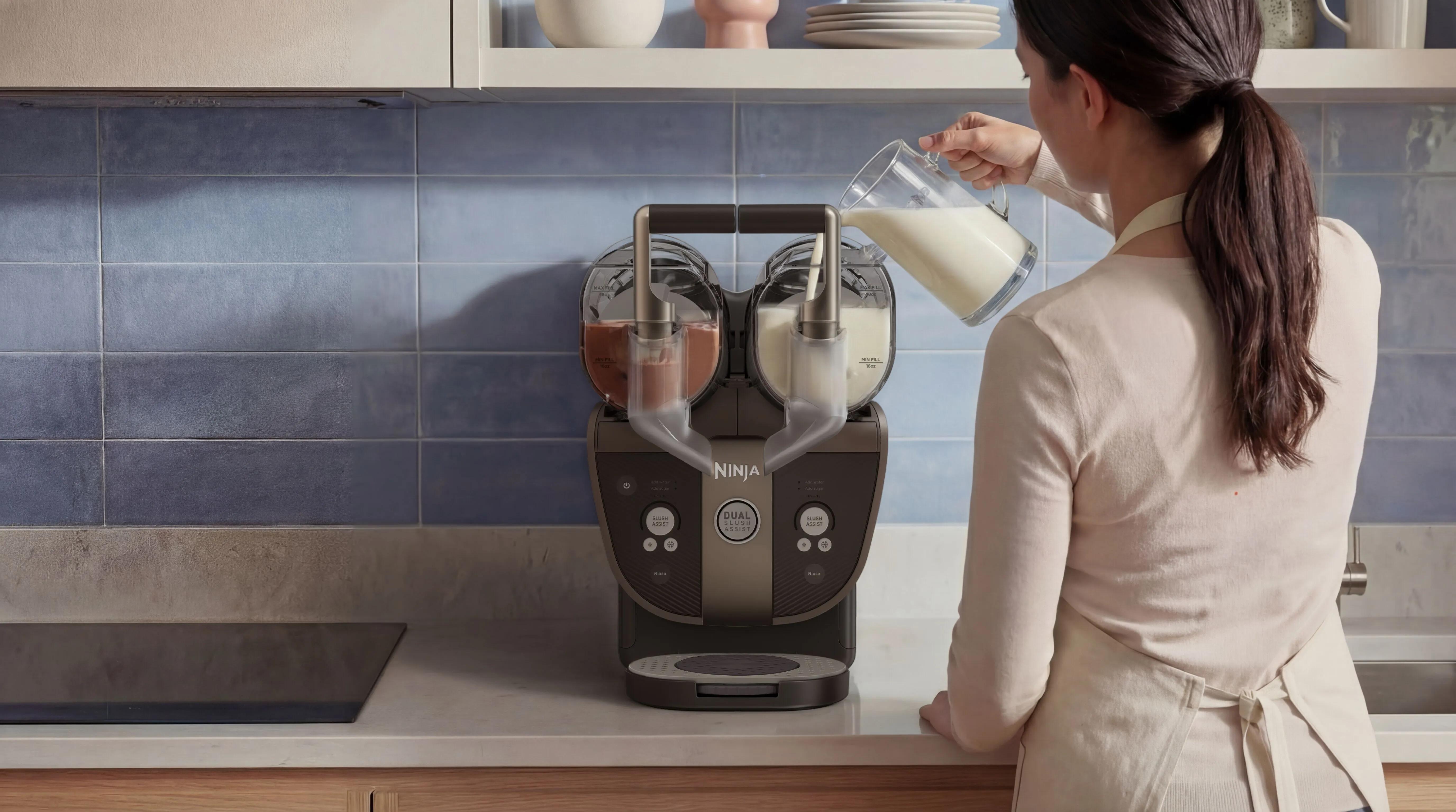 Eine Frau gießt Milch in einen doppelten Ninja-Slush-Maker in einer Küche mit blauen Fliesen.