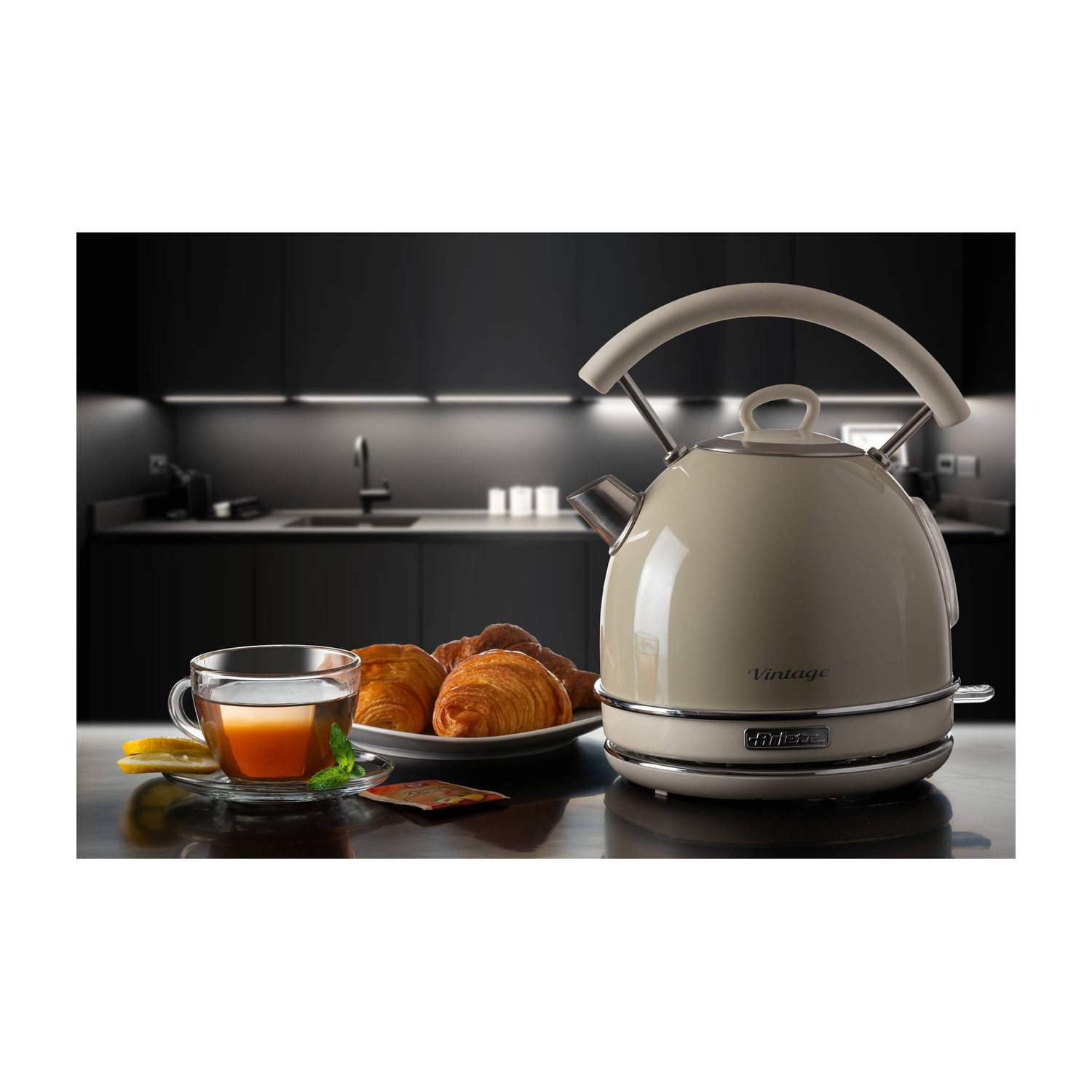 Bollitore vintage beige con tazza di tè, limoni e croissant su un bancone scuro.