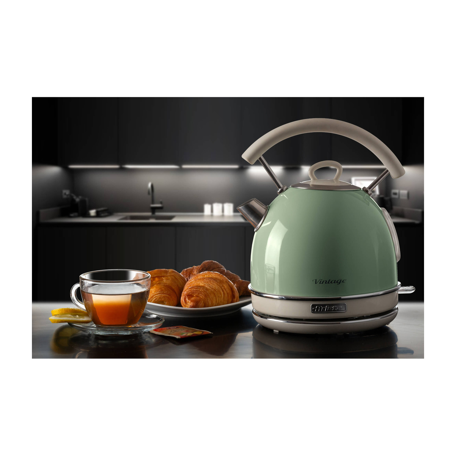 Un bollitore vintage verde pastello, una tazza di tè e croissant su un bancone con cucina sfocata sullo sfondo.