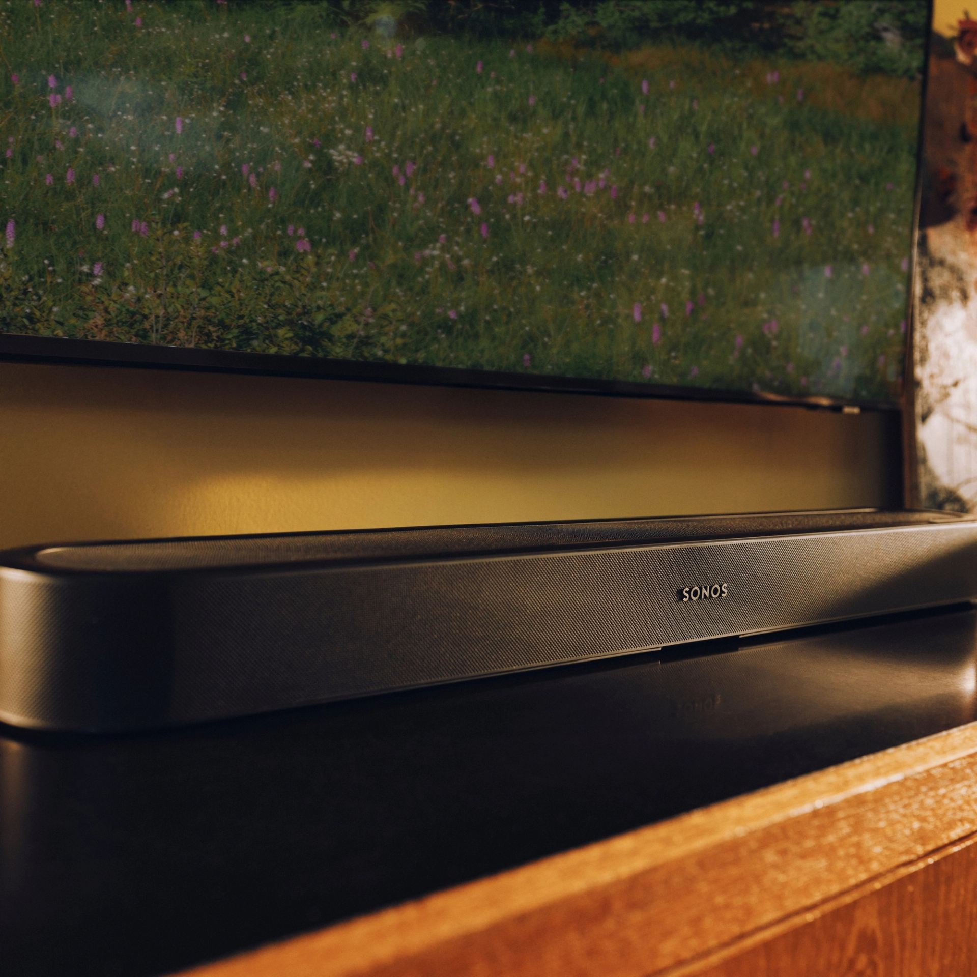 Egy fekete Sonos hangprojektor áll egy sötét felületen egy TV képernyő előtt.