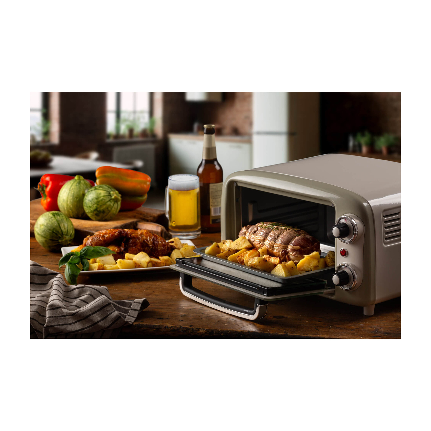 Un mini forno beige su un tavolo di legno con carne, patate, birra e verdure sullo sfondo.