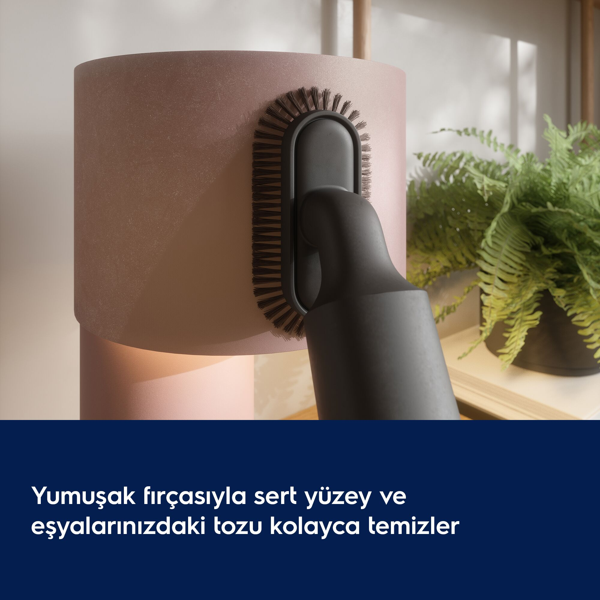 Siyah elektrik süpürgesi fırçası pembe abajuru temizliyor. Arkada yeşil saksı bitkisi var.