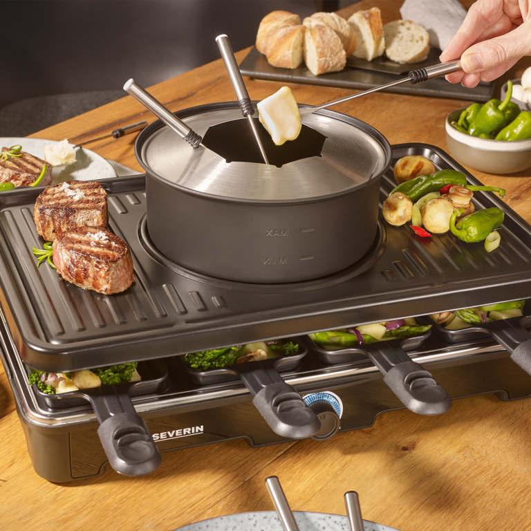 Tischgrill mit Raclette, Fleisch auf dem oberen Grill und Gemüse in kleinen Pfännchen.
