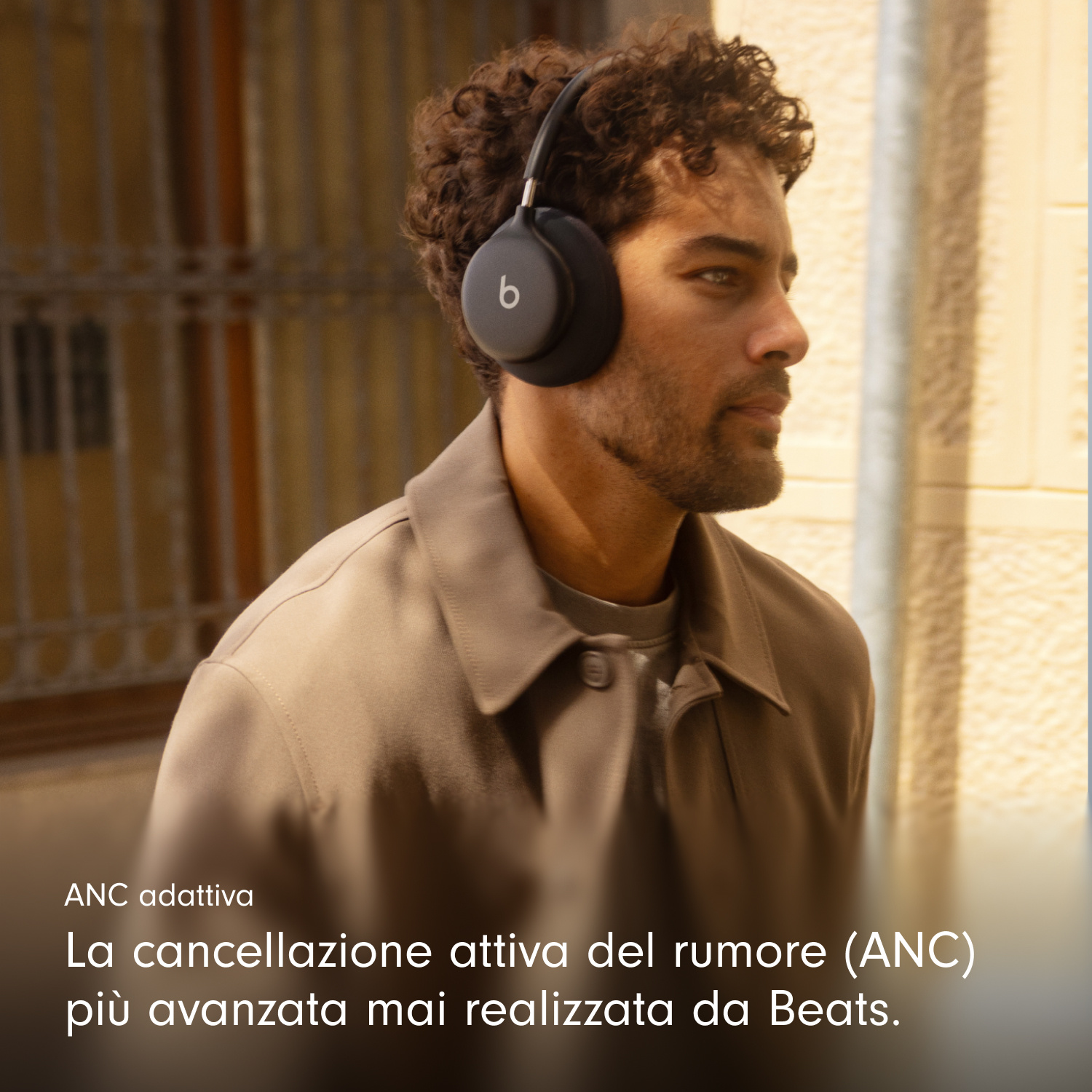 Un uomo con capelli scuri ricci indossa cuffie Beats nere e una giacca beige all'aperto.
