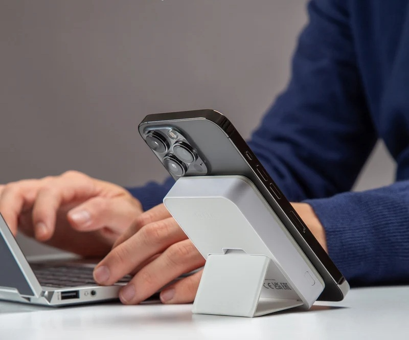Smartfon na białej podstawce obok laptopa, dłonie w tle.