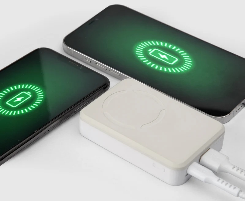 Dwa smartfony obok białego powerbanka podłączonego dwoma kablami, z wyświetlającą się na ekranie ikoną ładowania.