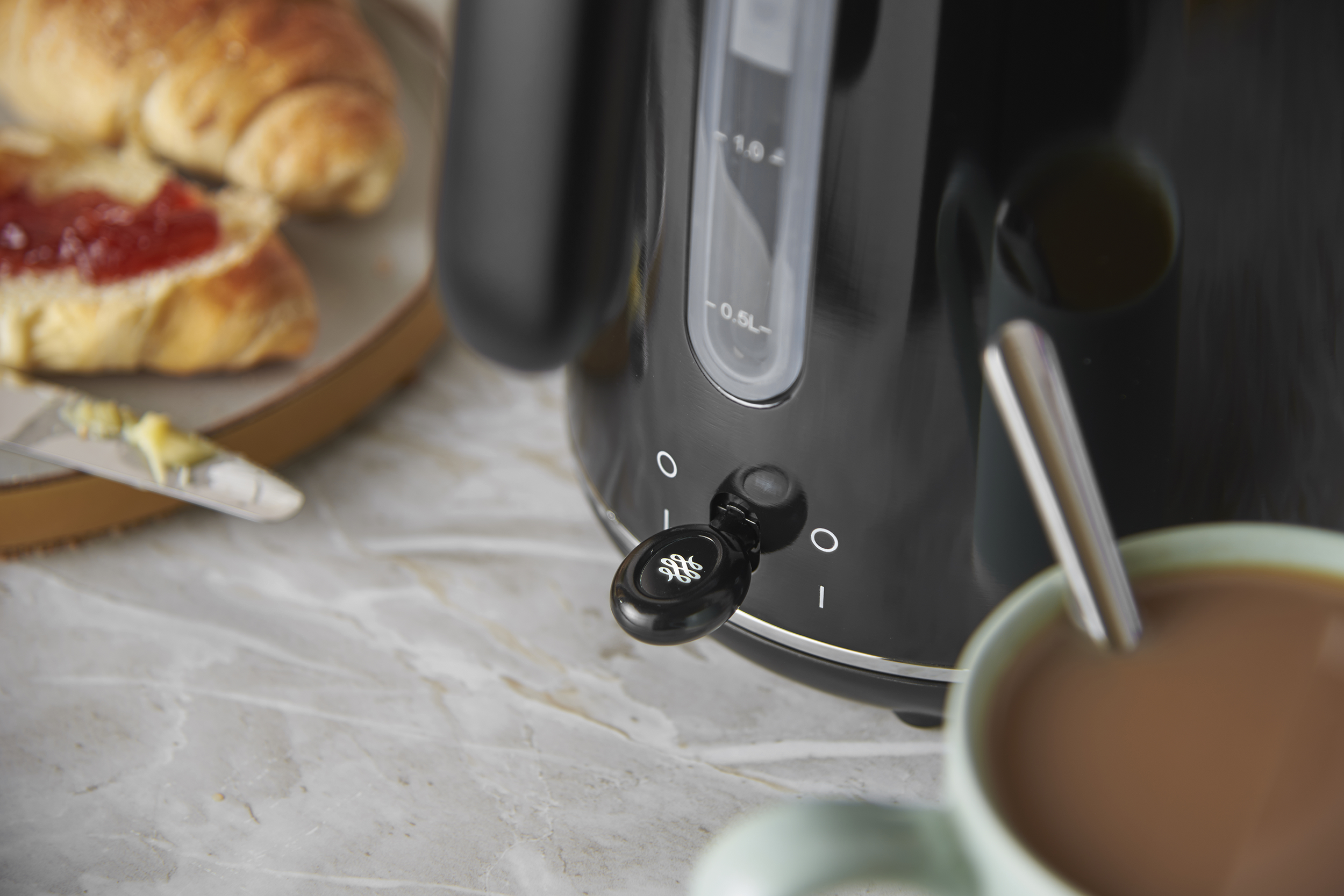 Egy fekete vízforraló áll a pulton lekváros croissant-ok és egy csésze kávé mellett.