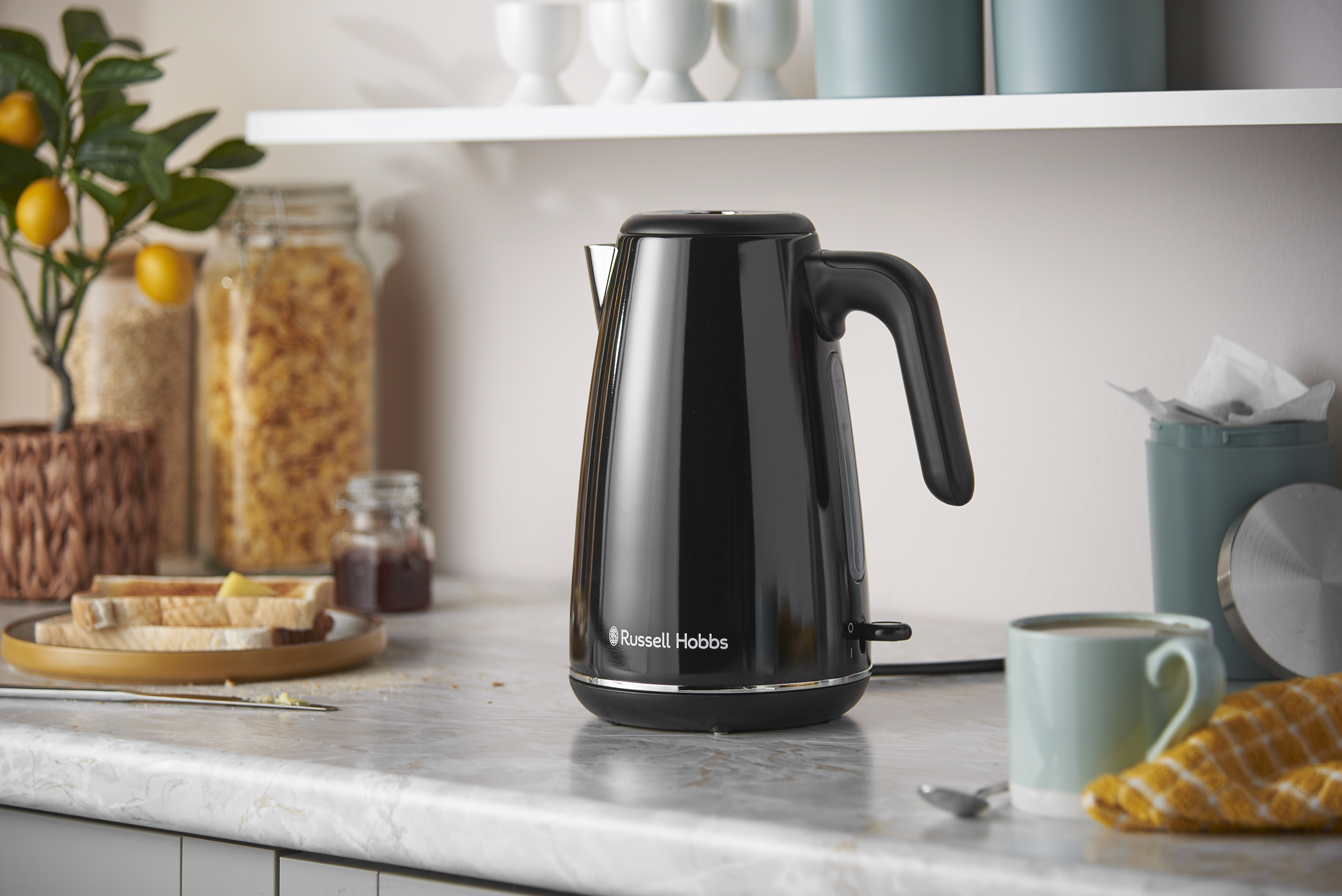 Egy fekete Russell Hobbs elektromos vízforraló márvány konyhapulton, pirítóssal, bögrével és üvegekkel a háttérben.
