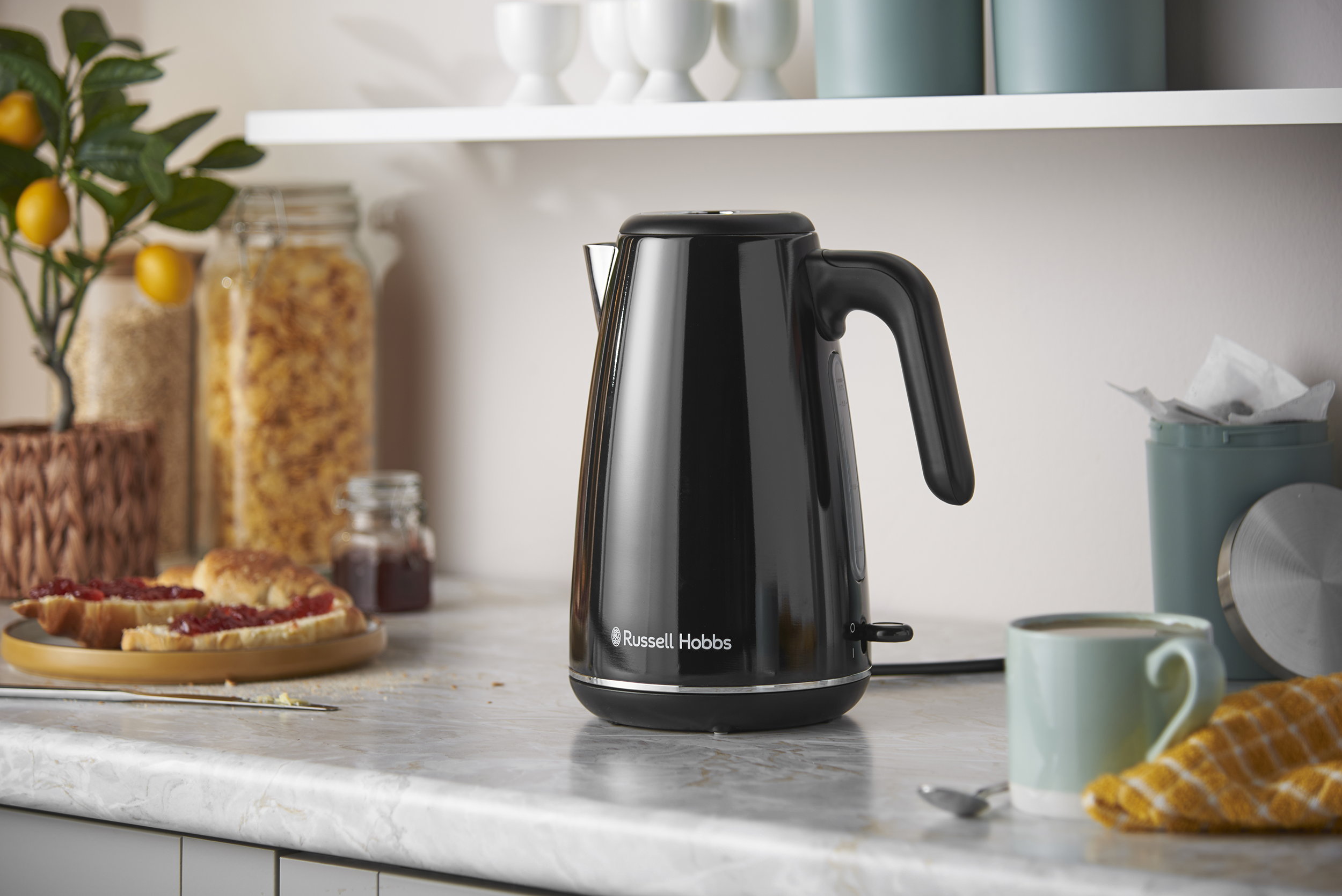 Egy fekete Russell Hobbs vízforraló áll egy márvány konyhai pulton, háttérben élelmiszerekkel.