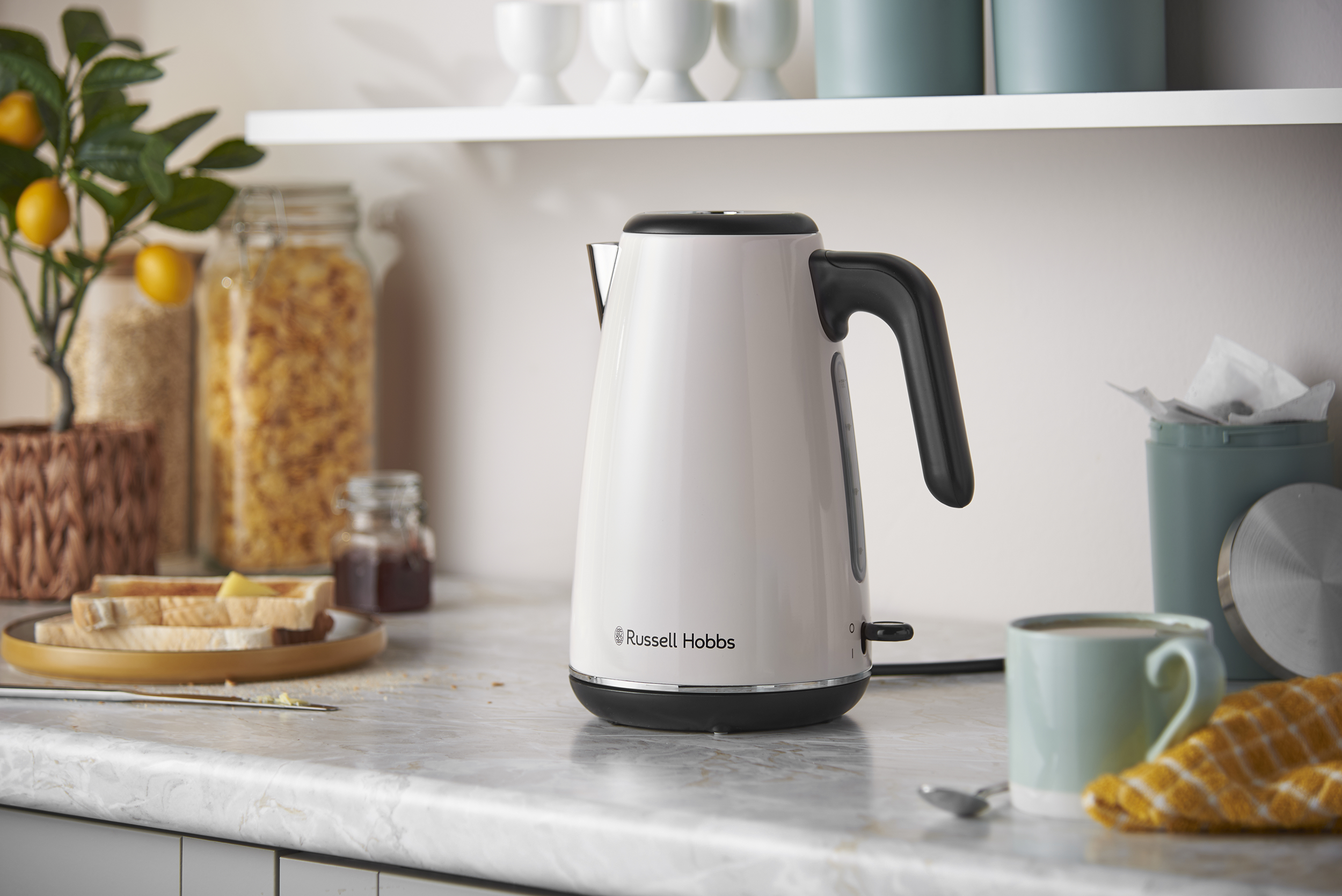Egy fehér Russell Hobbs vízforraló márvány munkalapon, pirítóssal, bögrével és kamraüvegekkel a háttérben.