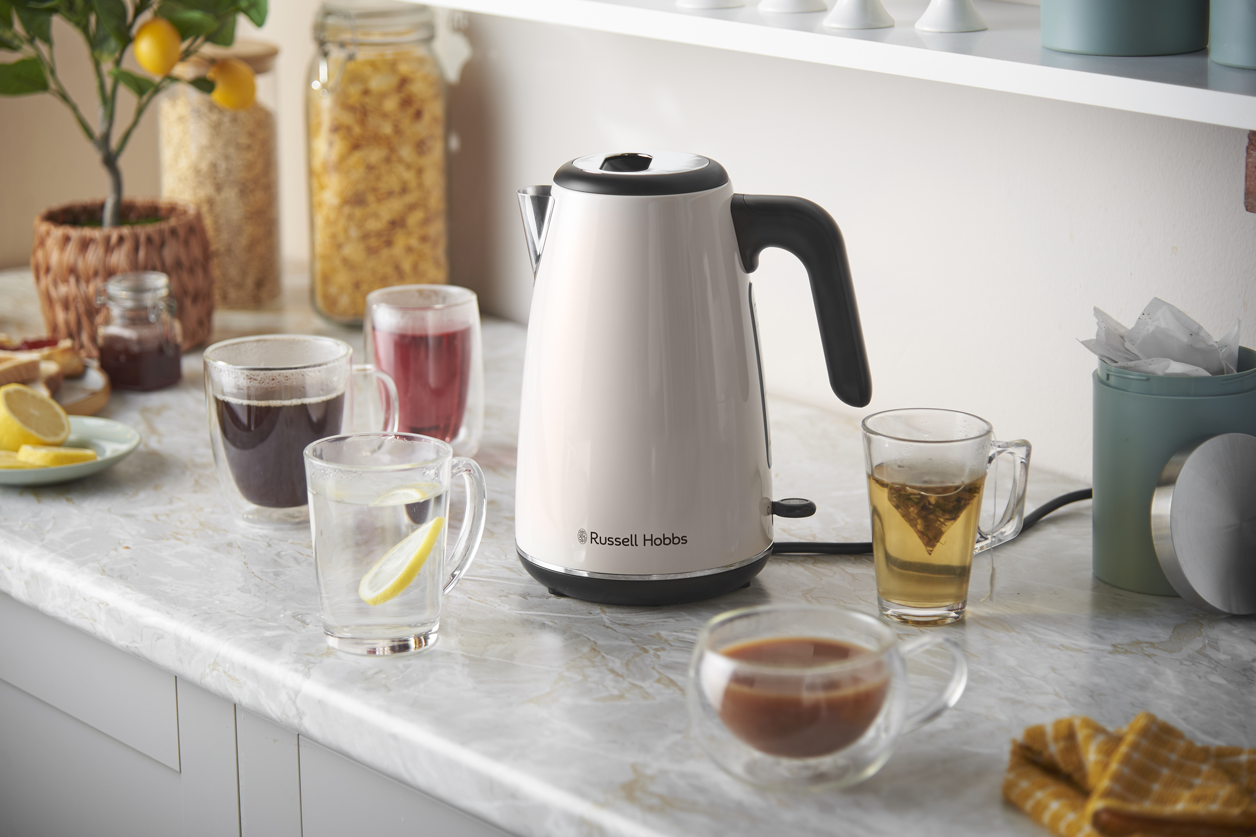Egy fehér Russell Hobbs vízforraló márvány munkalapon, italos poharakkal és élelmiszerekkel körülvéve.