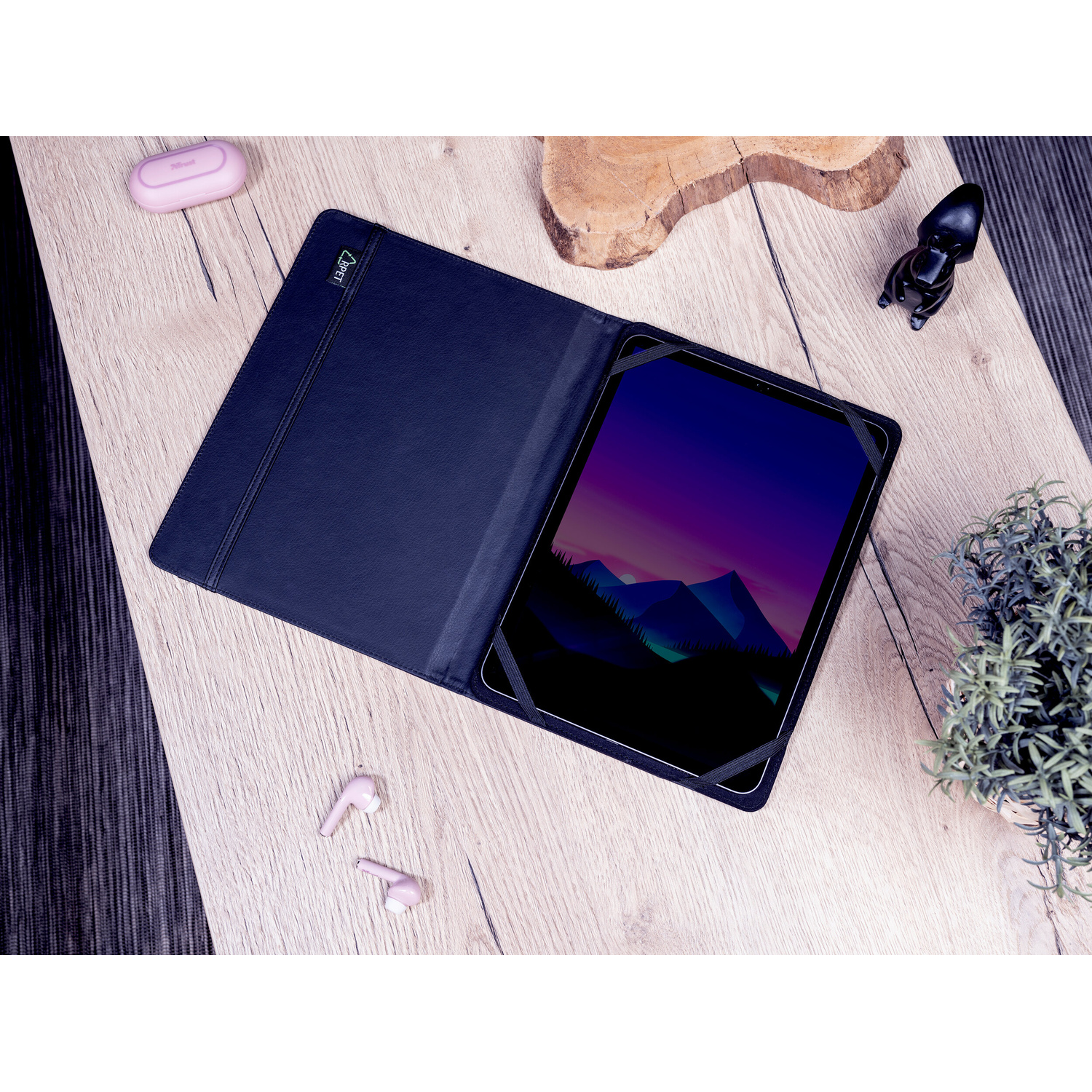 Un tablet in custodia blu scuro, auricolari rosa, una pianta e una piccola statuetta su una scrivania in legno.