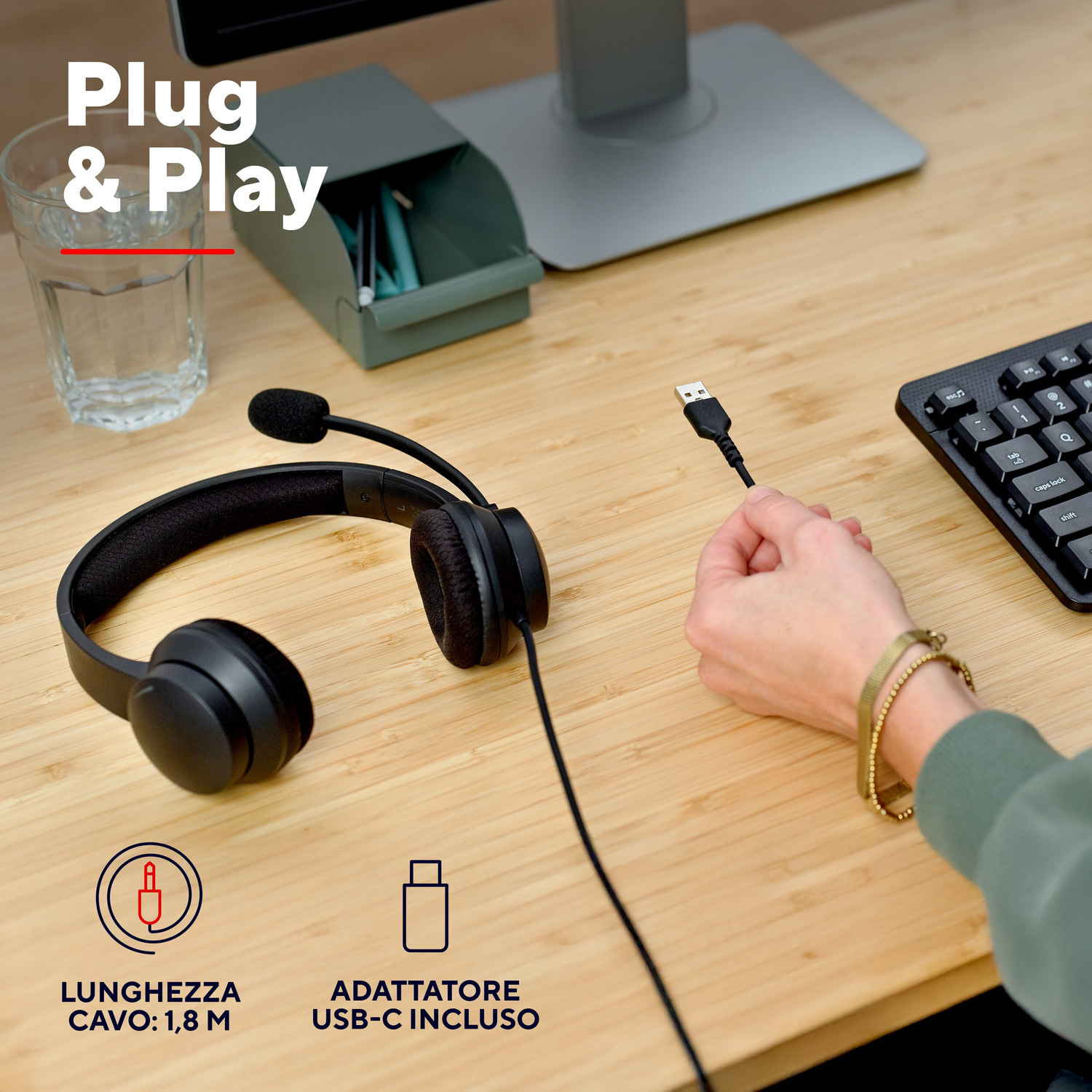 Mano regge il cavo USB di cuffie nere con microfono su scrivania in legno vicino alla tastiera.