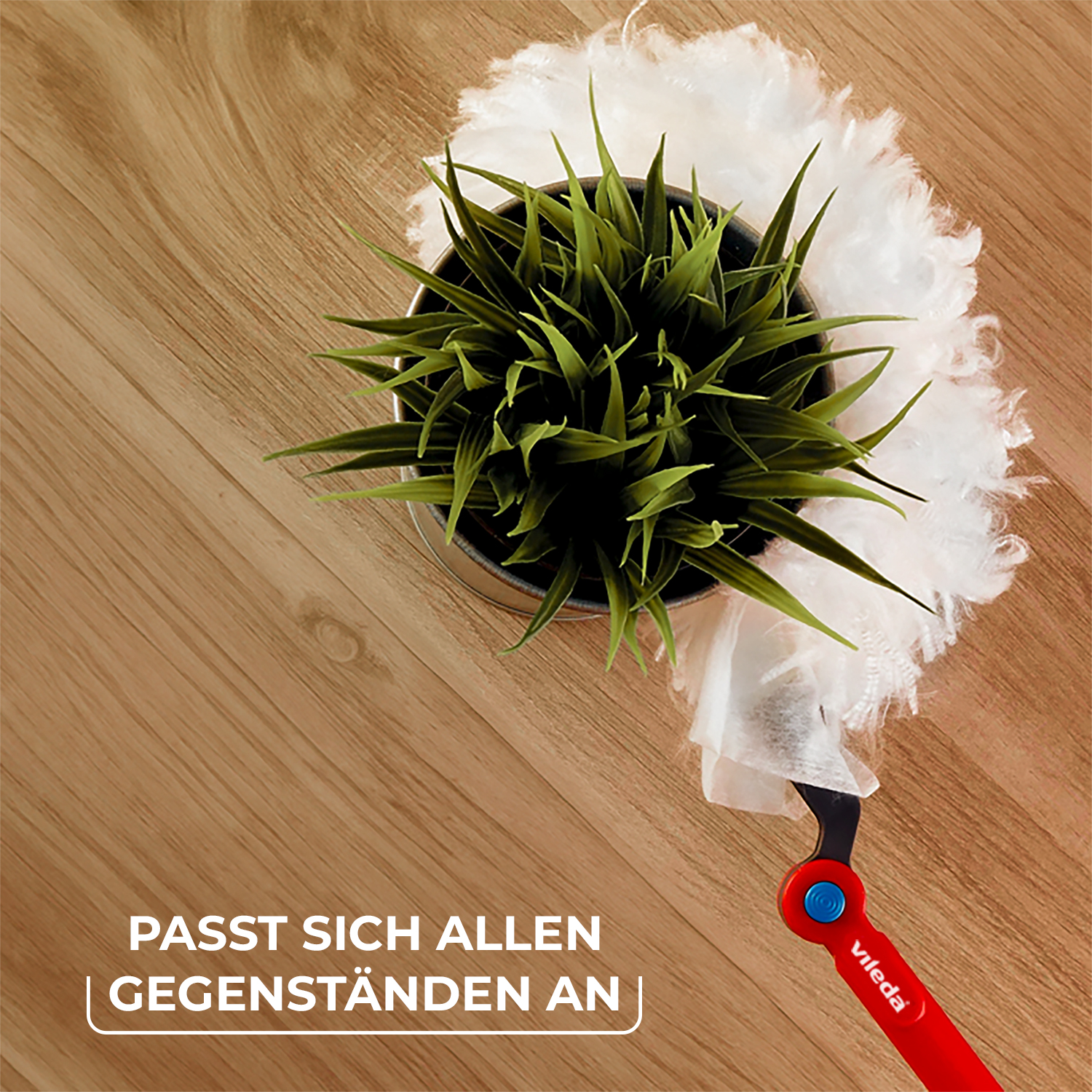 Ein weißer Staubwedel reinigt einen grünen Topf mit Gras auf einer Holzoberfläche.