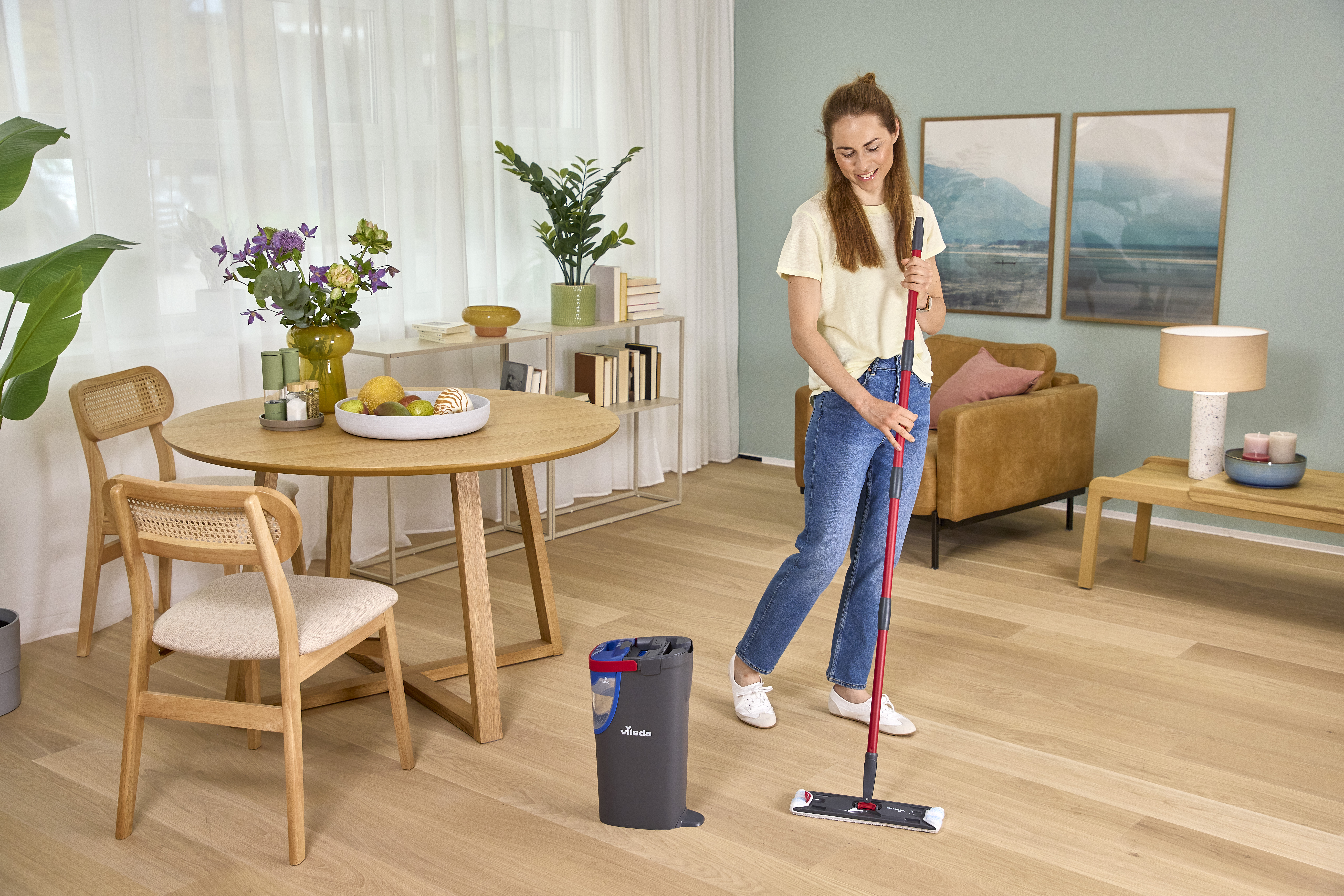 Eine Frau wischt den Holzboden mit einem roten Wischmopp in einem hellen, gemütlichen Wohnzimmer.