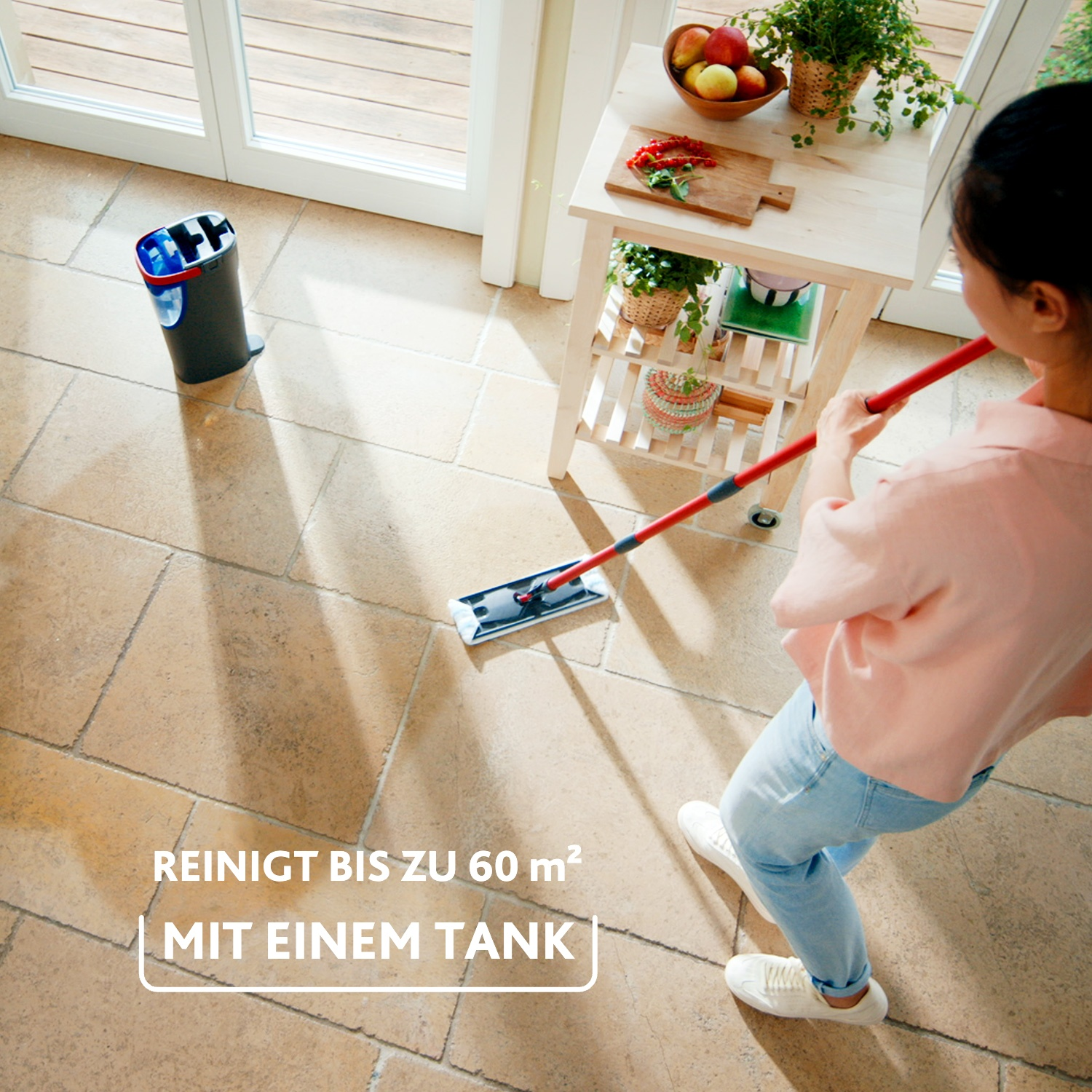 Eine Frau wischt einen Fliesenboden mit einem roten Wischmop; daneben steht ein Eimer und eine Holzkommode.
