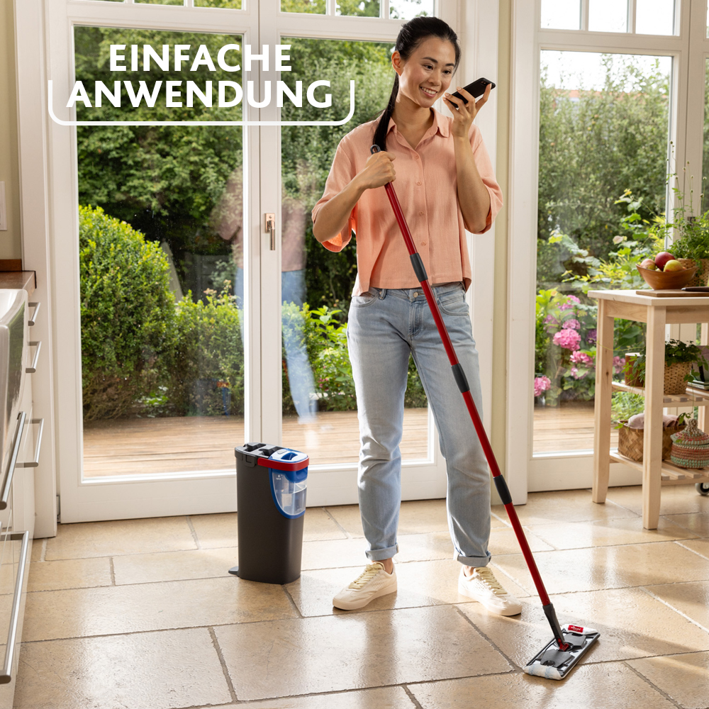 Eine lächelnde Frau wisch den Boden und telefoniert, mit einem Eimer daneben und Blick in den Garten.