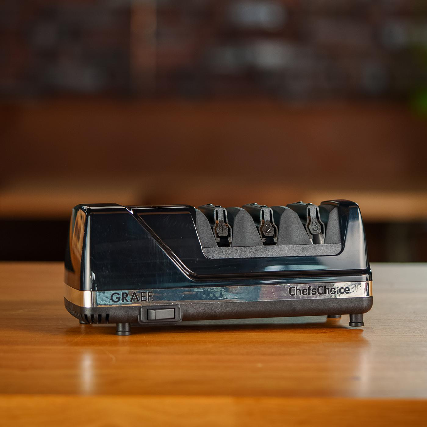 Ein schwarzer elektrischer Messerschärfer mit der Aufschrift Graef und Chef's Choice auf einer Holzoberfläche.