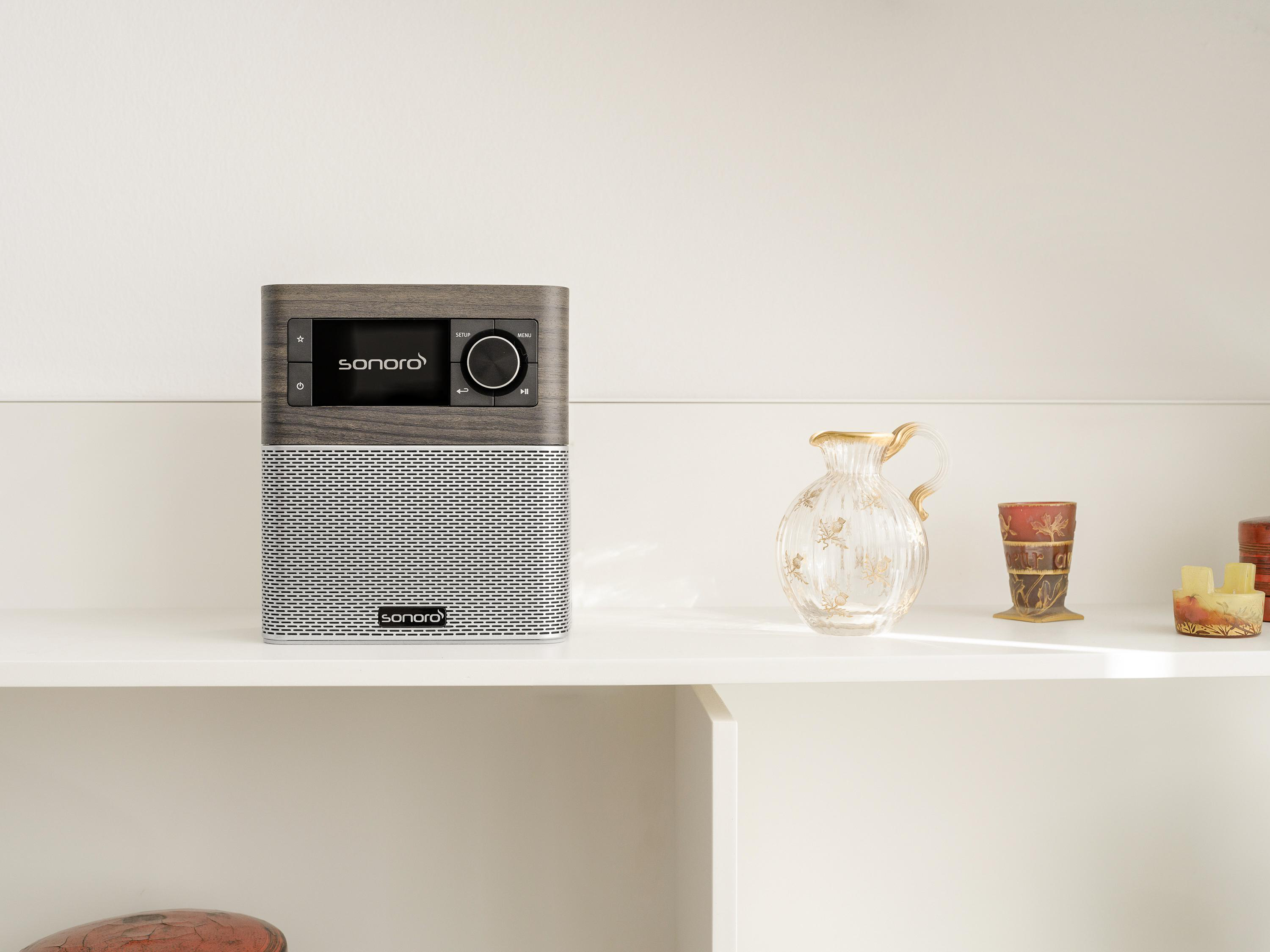 Ein Sonoro-Radio mit Holzfront auf einem weißen Regal neben dekorativen Glas- und Keramikartikeln.
