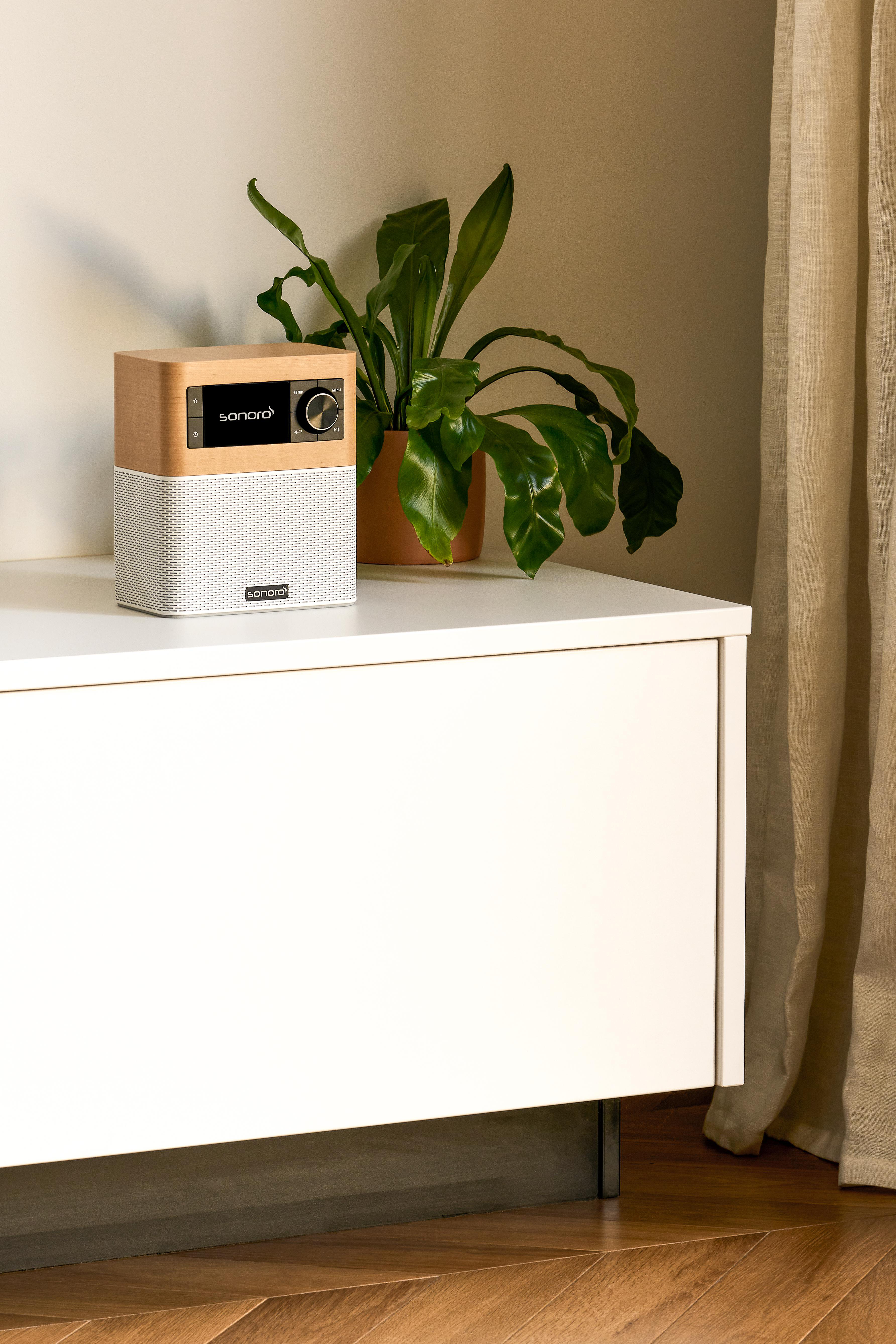 Ein hölzernes Sonoro-Radio steht auf einem weißen Sideboard neben einer Topfpflanze, vor einer beigen Wand und einem Vorhang.