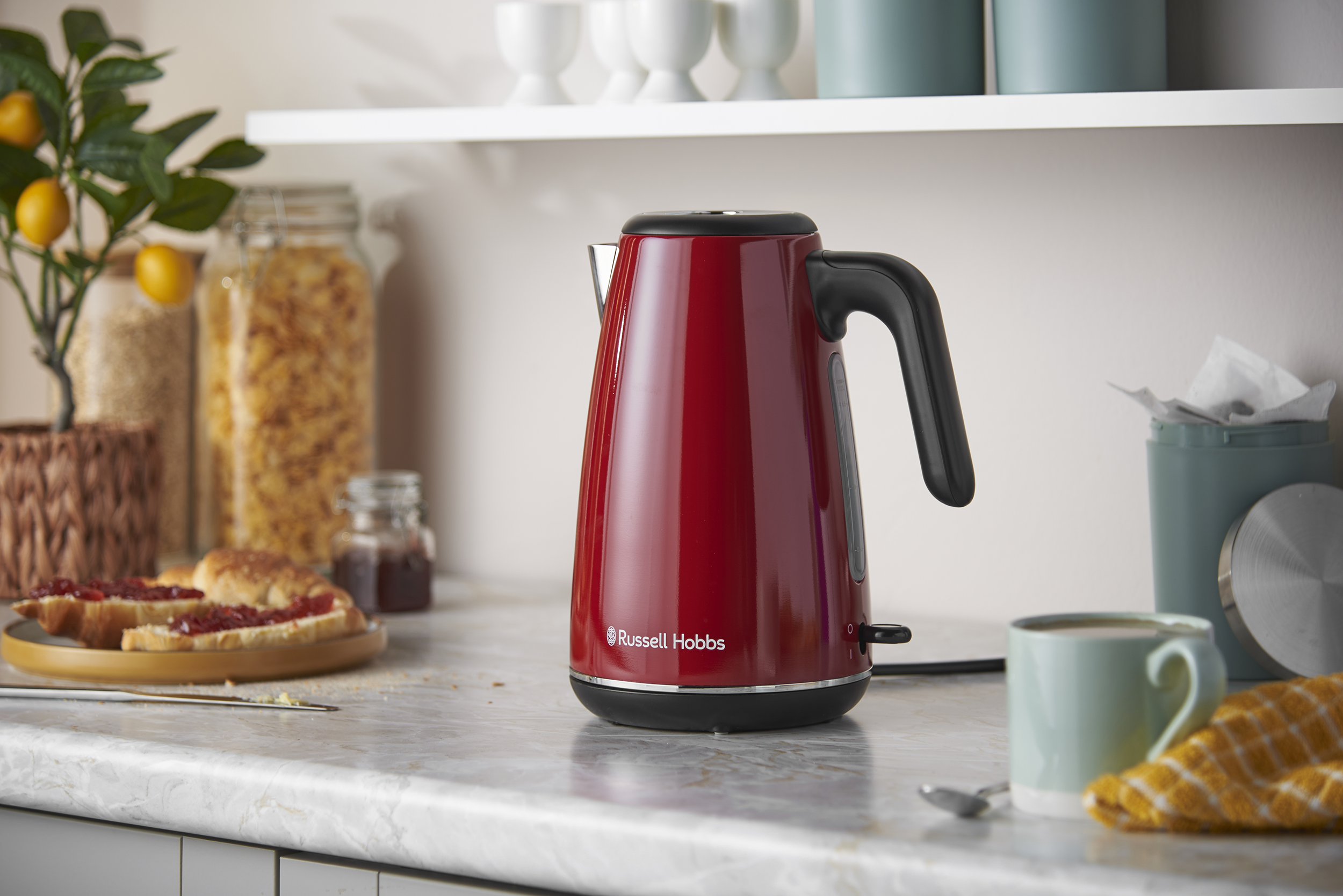 Egy piros Russell Hobbs vízforraló márvány pulton egy konyhában, háttérben élelmiszerekkel.