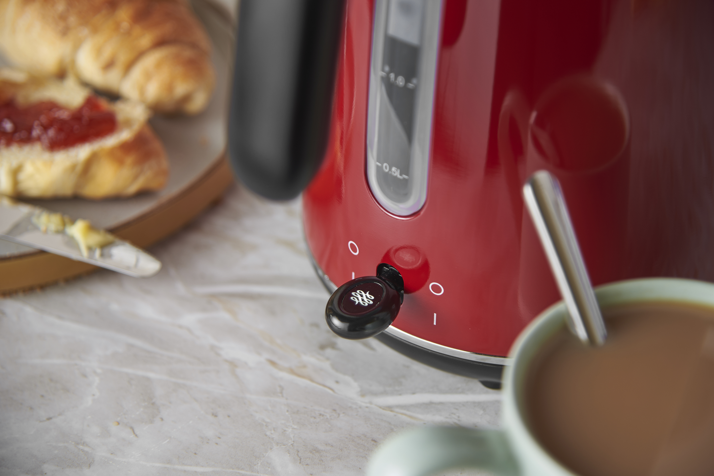 Fényes piros elektromos vízforraló márvány pulton, egy csésze kávéval és croissant-nal a háttérben.
