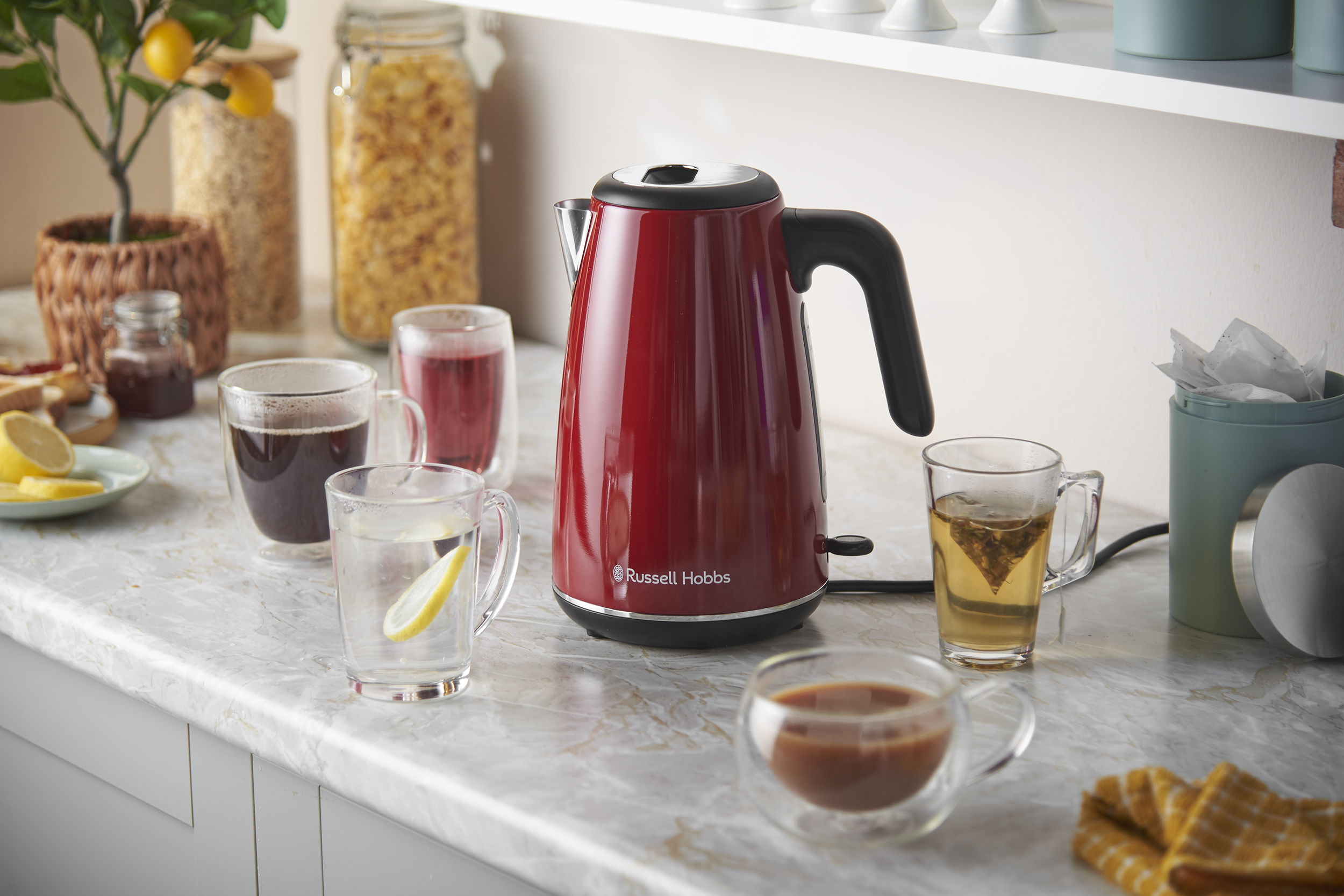 Piros Russell Hobbs vízforraló márvány munkalapon, körbevéve italos poharakkal és élelmiszertárolókkal.