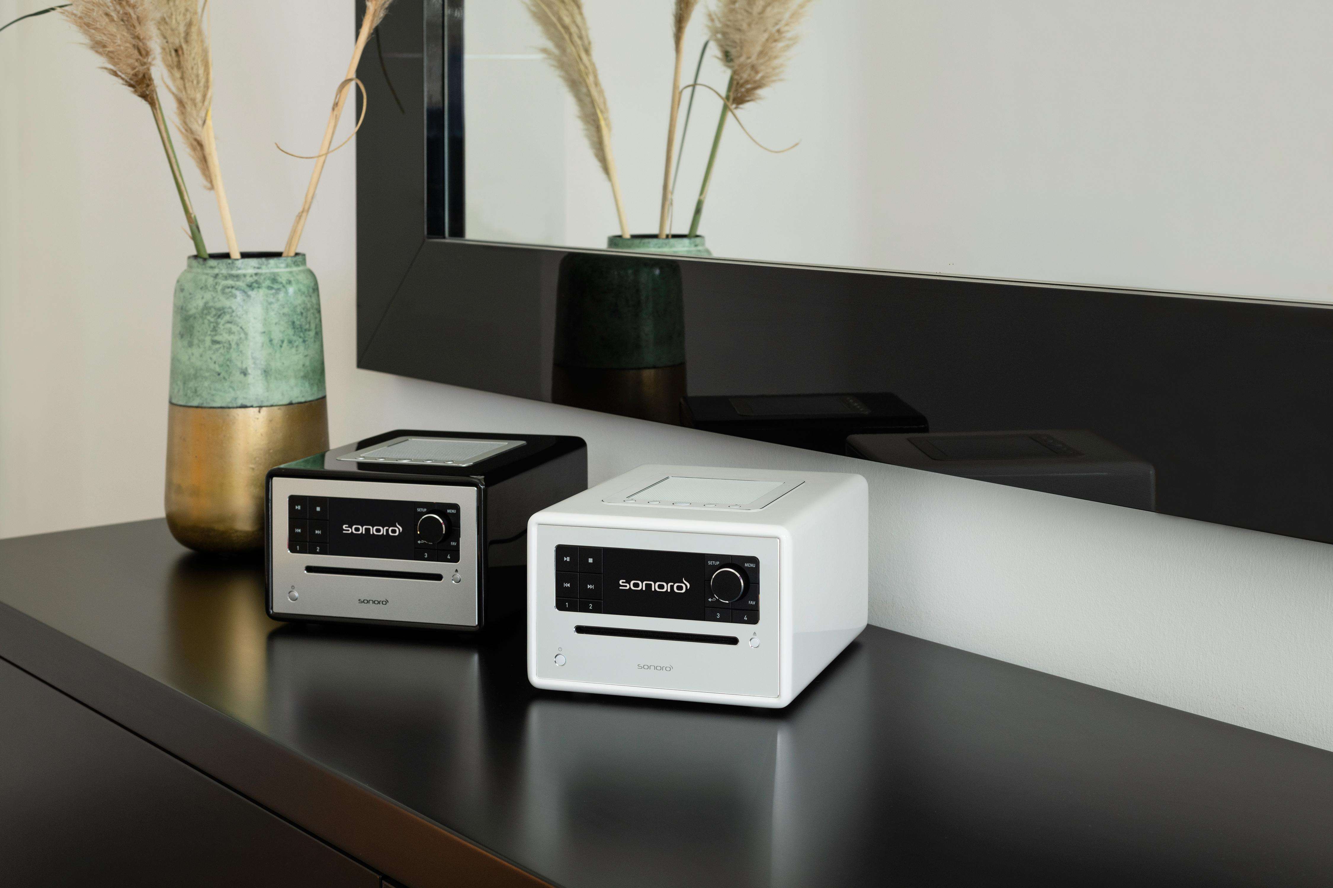 Zwei sonoro Musiksysteme, ein schwarzes und ein weißes, stehen auf einem dunklen Sideboard mit Vase und Spiegel.