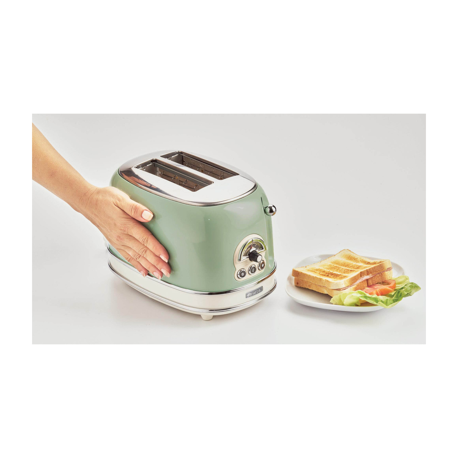 Una mano tocca un toaster verde chiaro, con un piatto di panini accanto su sfondo bianco.
