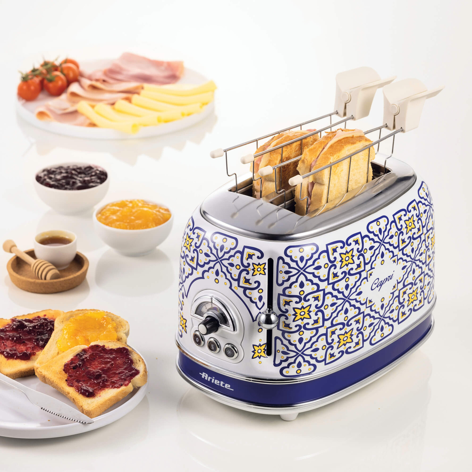Un tostapane blu e bianco con fantasia Capri e panini si trova su un tavolo con pane, marmettate e salumi.