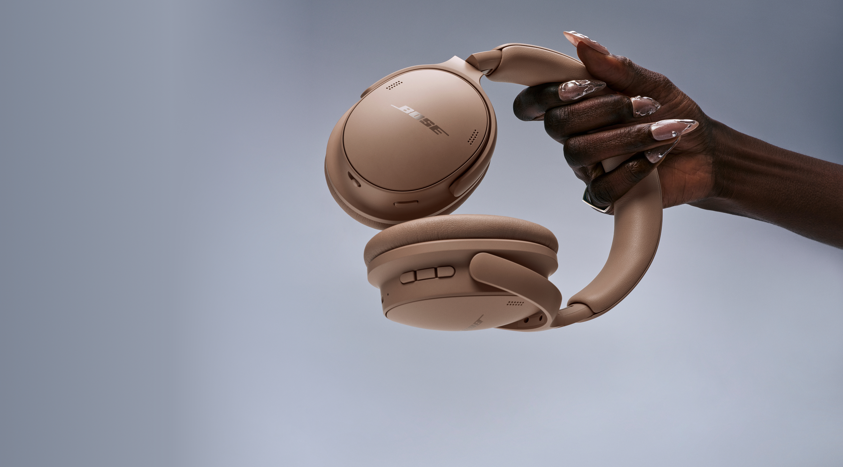 Eine Hand hält beige Bose-Kopfhörer vor einem grauen Hintergrund.