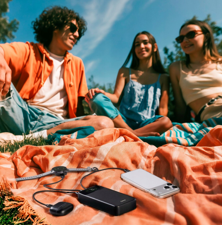 Trójka przyjaciół siedzi w parku, podczas gdy powerbank ładuje smartfon, zegarek i słuchawki.
