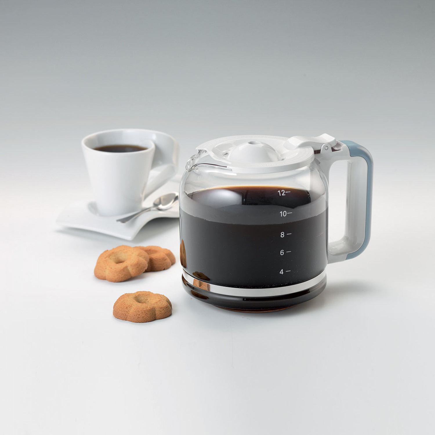 Una caffettiera piena con scala graduata, una tazza di caffè e tre biscotti su sfondo bianco.