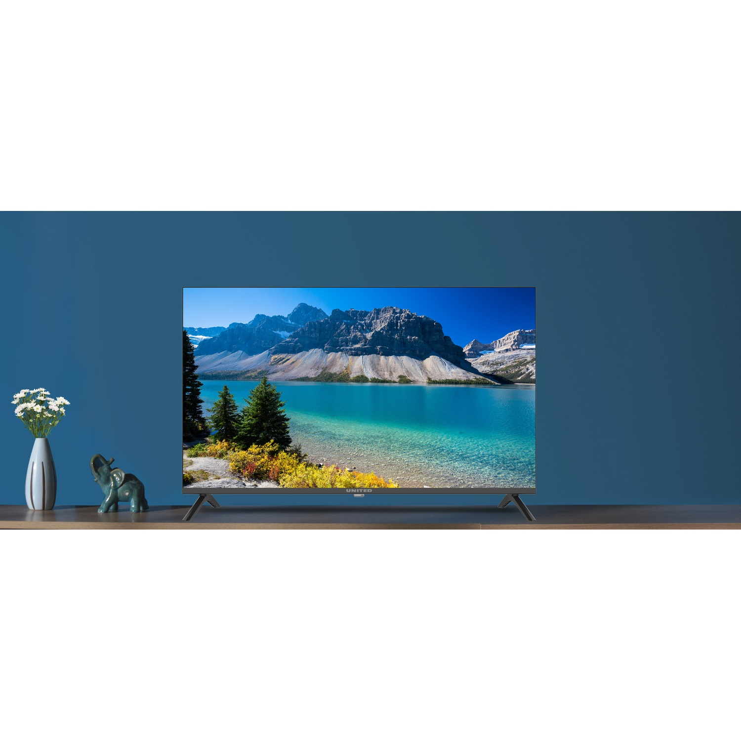 Una TV con paesaggio montano su scaffale di legno contro parete blu, affiancata da vaso e statuetta di elefante.