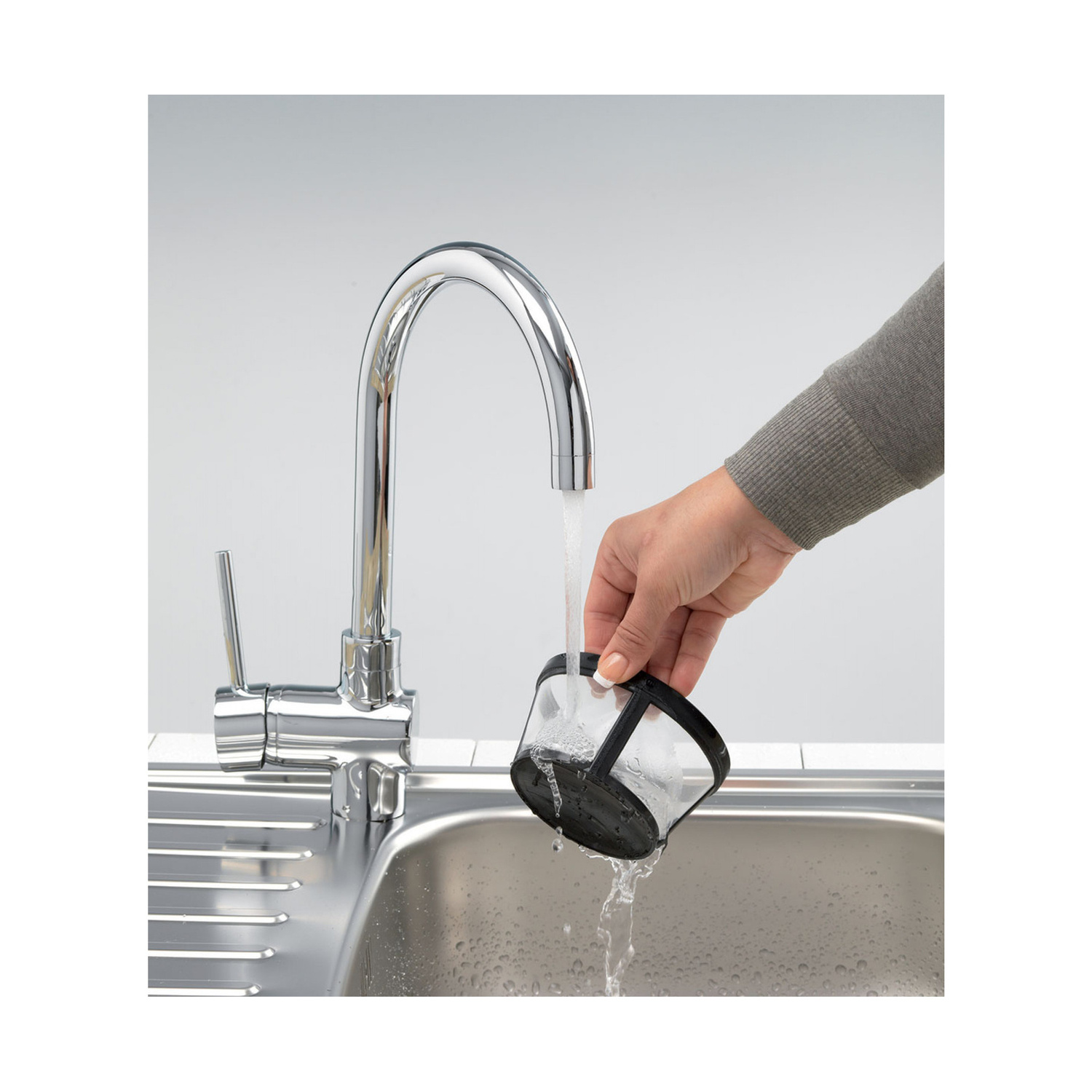 Una mano risciacqua un piccolo filtro nero sotto un moderno rubinetto cromato sopra un lavello in acciaio inossidabile.