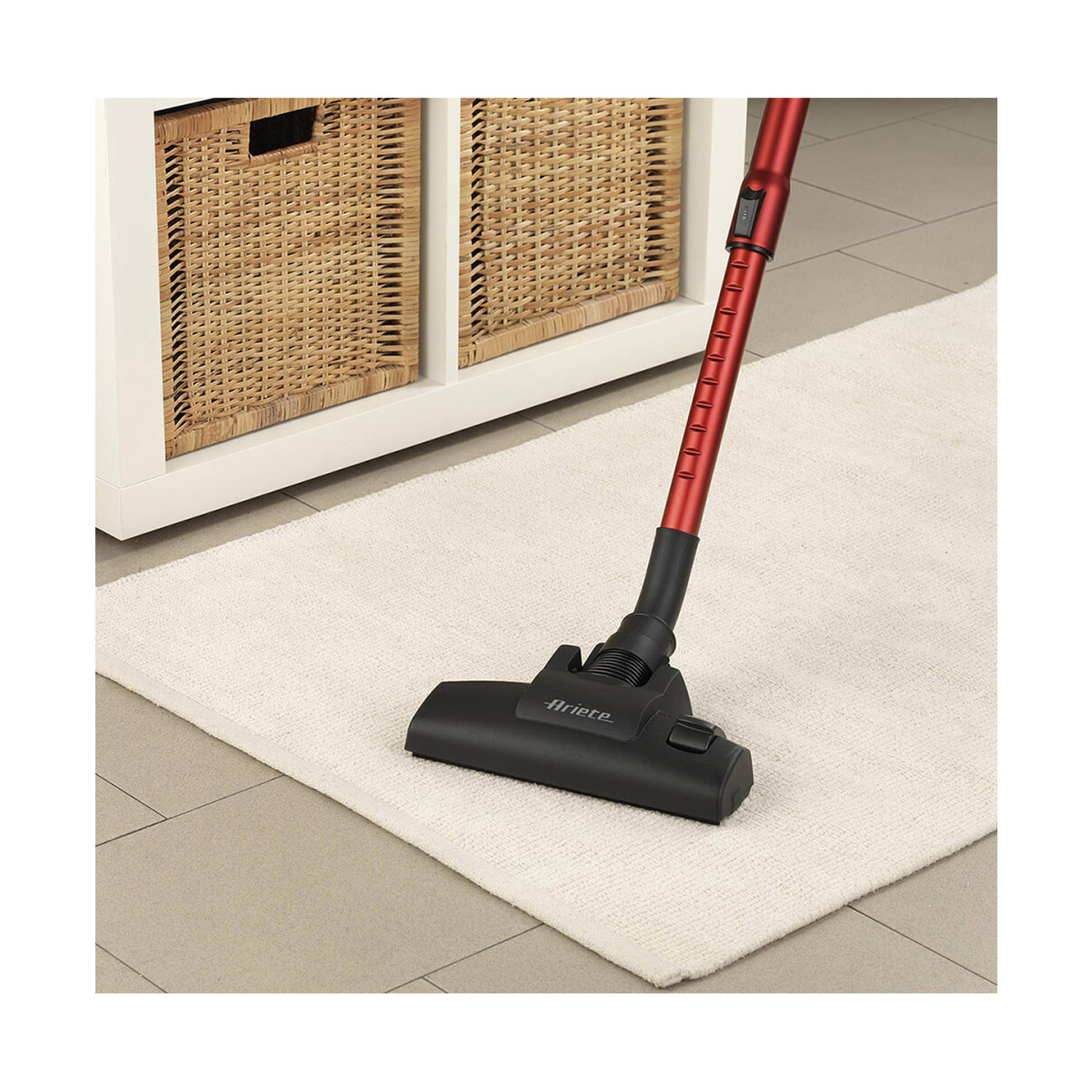 Un aspirapolvere Ariete rosso e nero aspira un tappeto bianco davanti a un mobile bianco con cesti in vimini.