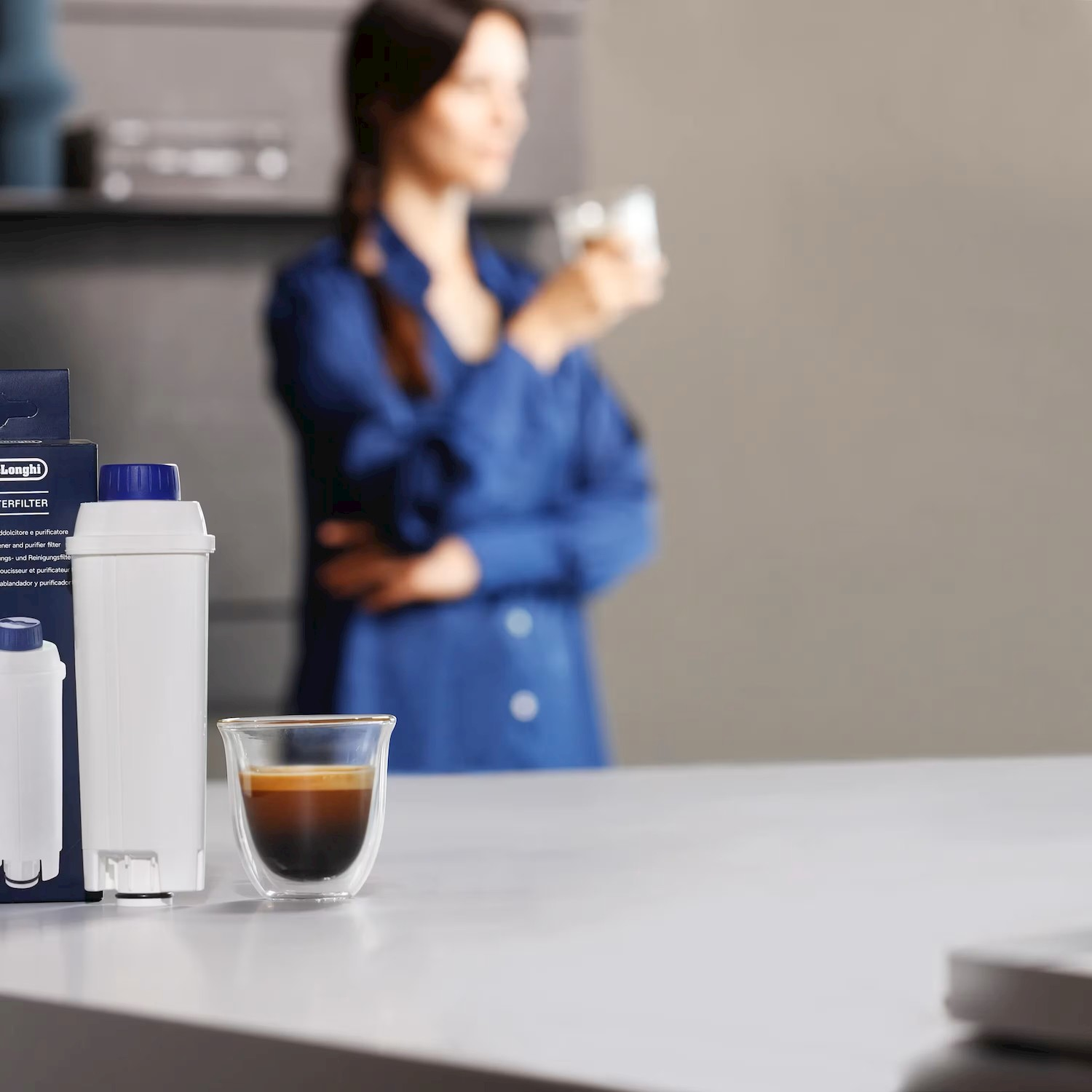 Un filtro per l'acqua e una tazzina da caffè sono su un bancone. Sfondo: una donna in blu beve caffè.