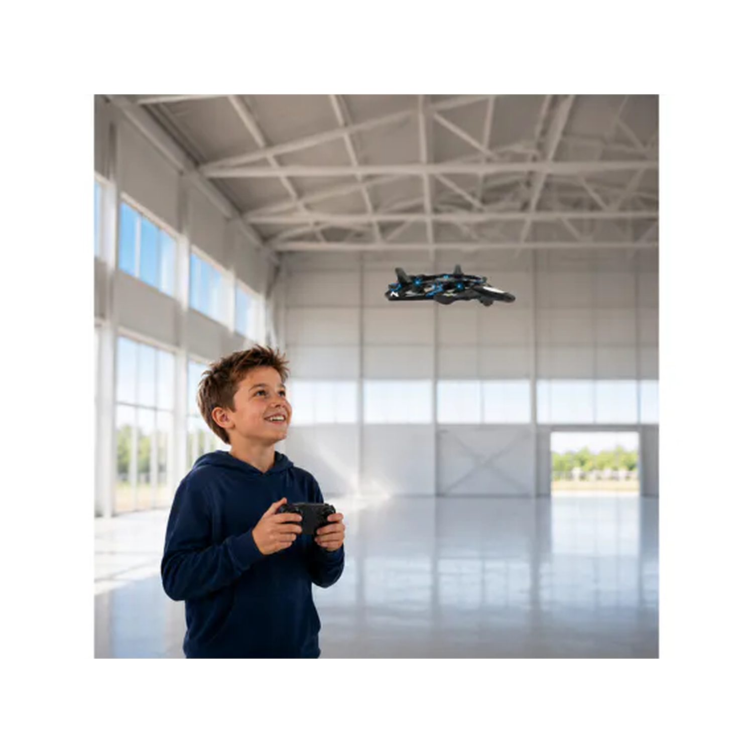 A smiling boy in a blue sweater controls a flying drone in a large, bright hall.