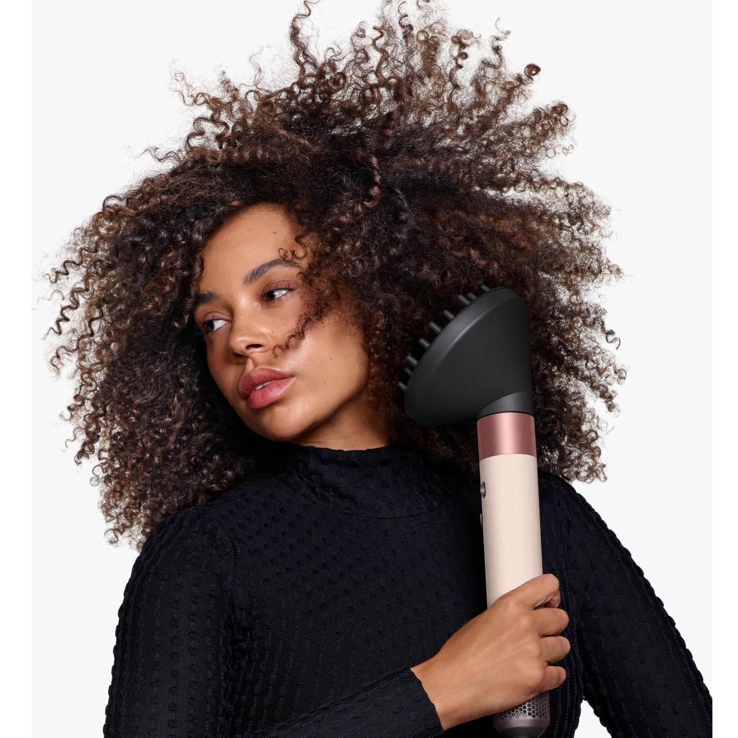 A woman with large, curly hair holds a hair styling tool against a white background.