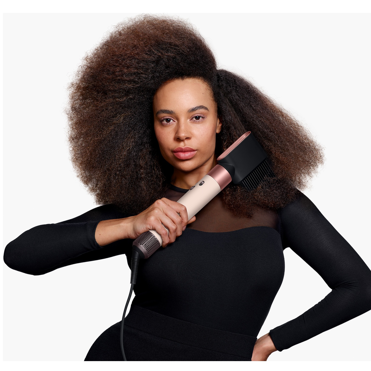 A woman with large afro hair holds a modern hair styling tool against a white background.