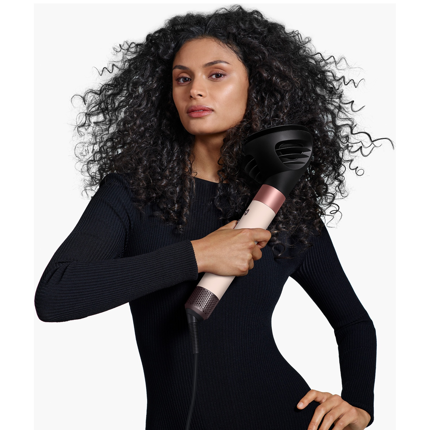 A woman with voluminous dark curly hair holds a pink hair dryer against a white background.
