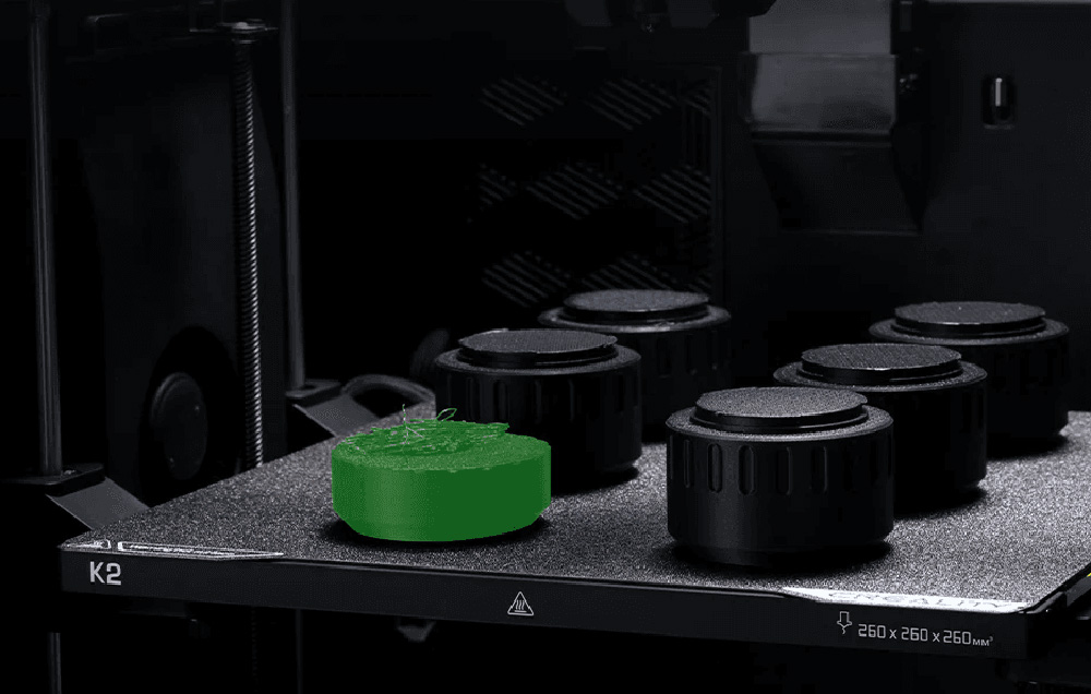 Egy 3D nyomtató zöld hengeres tárgyat nyomtat több fekete alkatrész mellett az építőlapon.