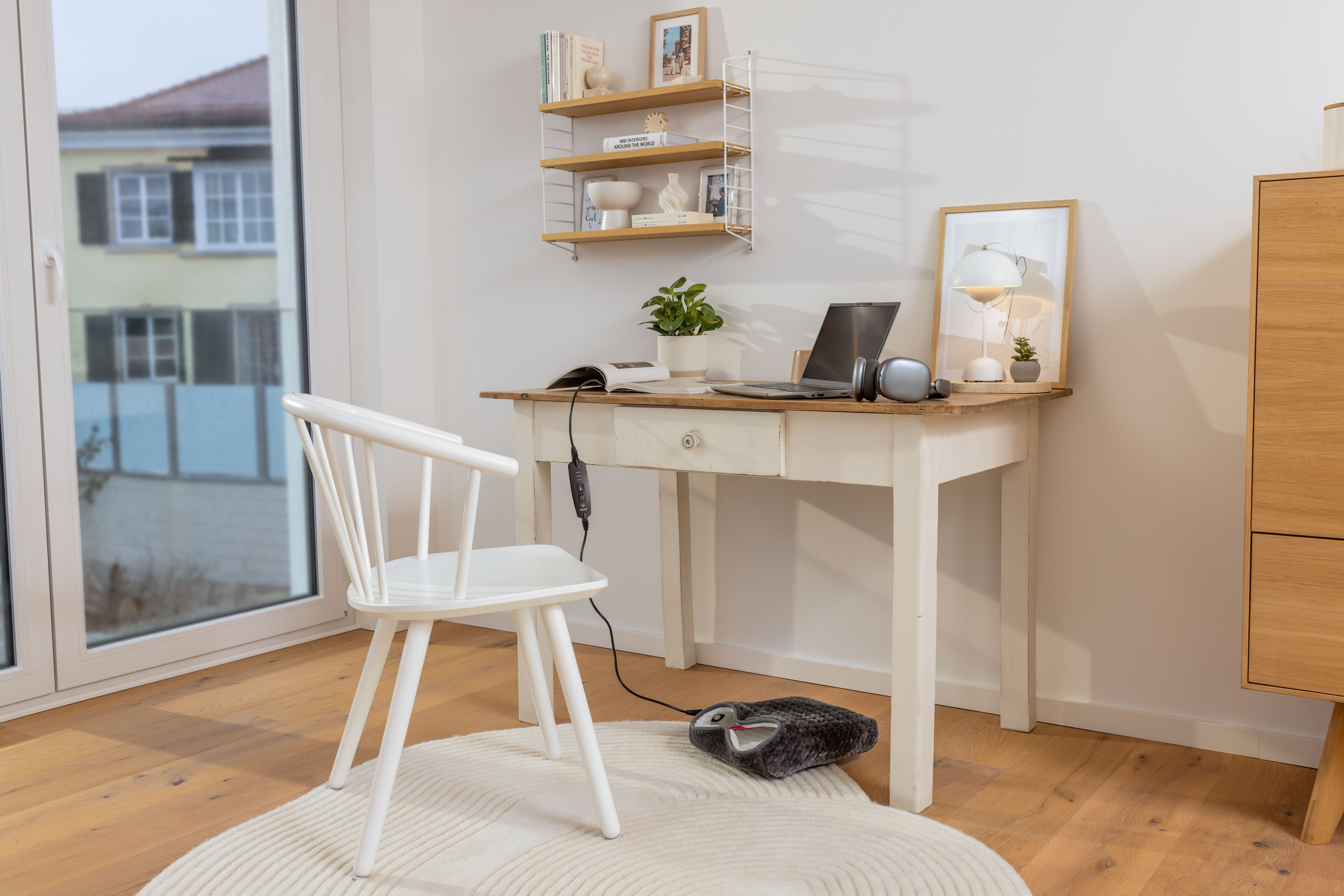 Ein weißer Holzschreibtisch mit Laptop, Lampe und Deko steht auf einem runden Teppich neben einer weißen Stuhl.