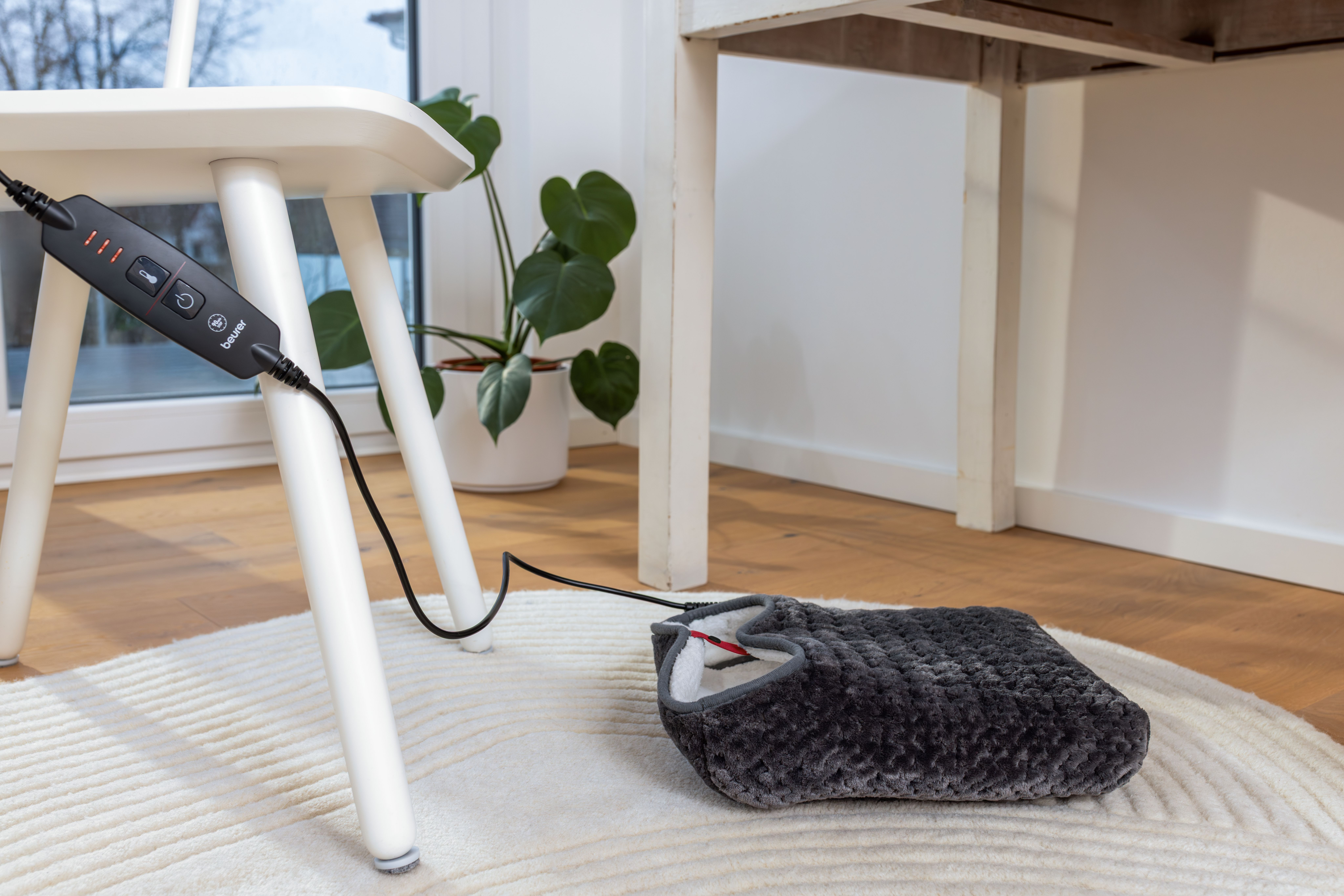 Ein grauer elektrischer Fußwärmer liegt auf einem beigen Teppich unter einem Tisch.