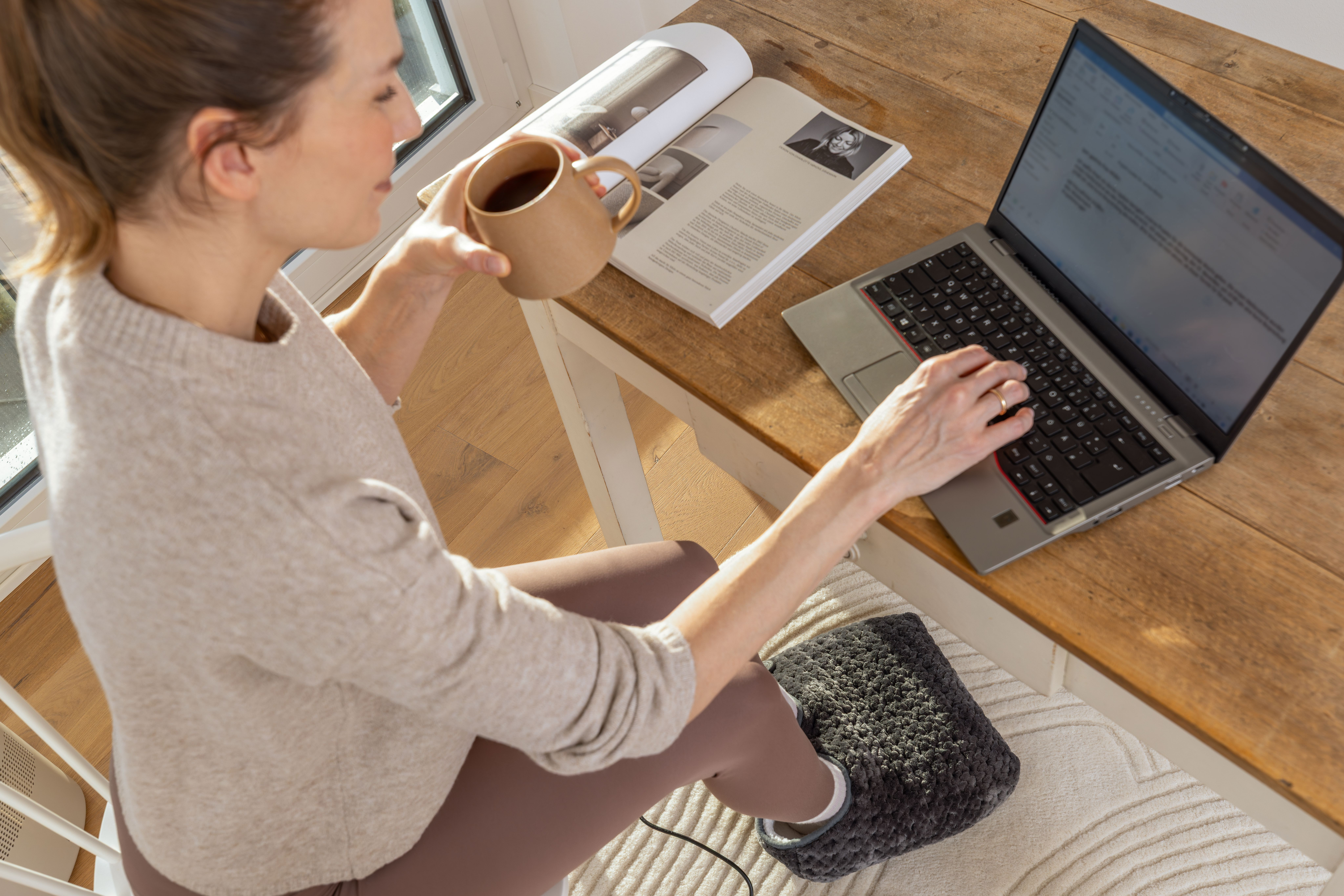 Eine Frau trinkt Kaffee und arbeitet am Laptop an einem Holztisch, ihre Füße ruhen auf einem wärmenden Fußwärmer.